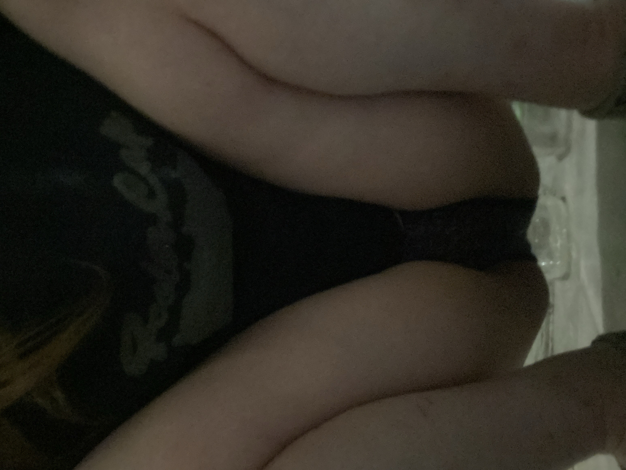 The image is a close-up, dimly lit shot of a person's legs and lower torso. The person is wearing dark underwear. The background is dark and out of focus, with some blurry white shapes that might be objects or a textured surface. There are no discernible location cues, time of day, or weather information. The image does not convey any specific emotion or activity other than the presentation of a body part. There is no visible text in the image.