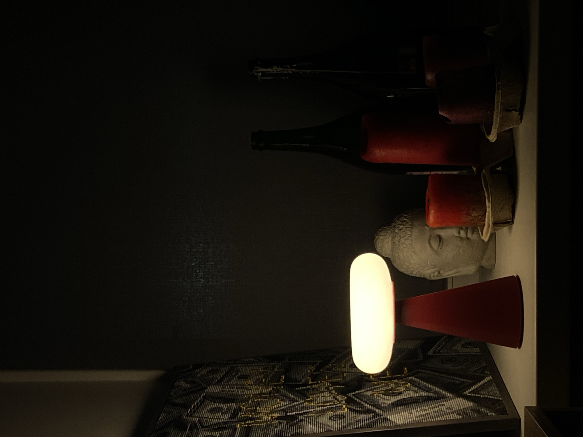 A dark, low-angle shot features a shelf containing a lamp, wine bottles, candles, and a bust. The lamp, with a white cylindrical light and a red conical base, sits on a patterned surface at the bottom of the frame. Above the lamp, a light gray bust is visible, positioned next to red candles in holders. Wine bottles lie horizontally along the shelf. The background is dark and indistinct. The location, Votkinsk, Russia, is not discernible from the image content.