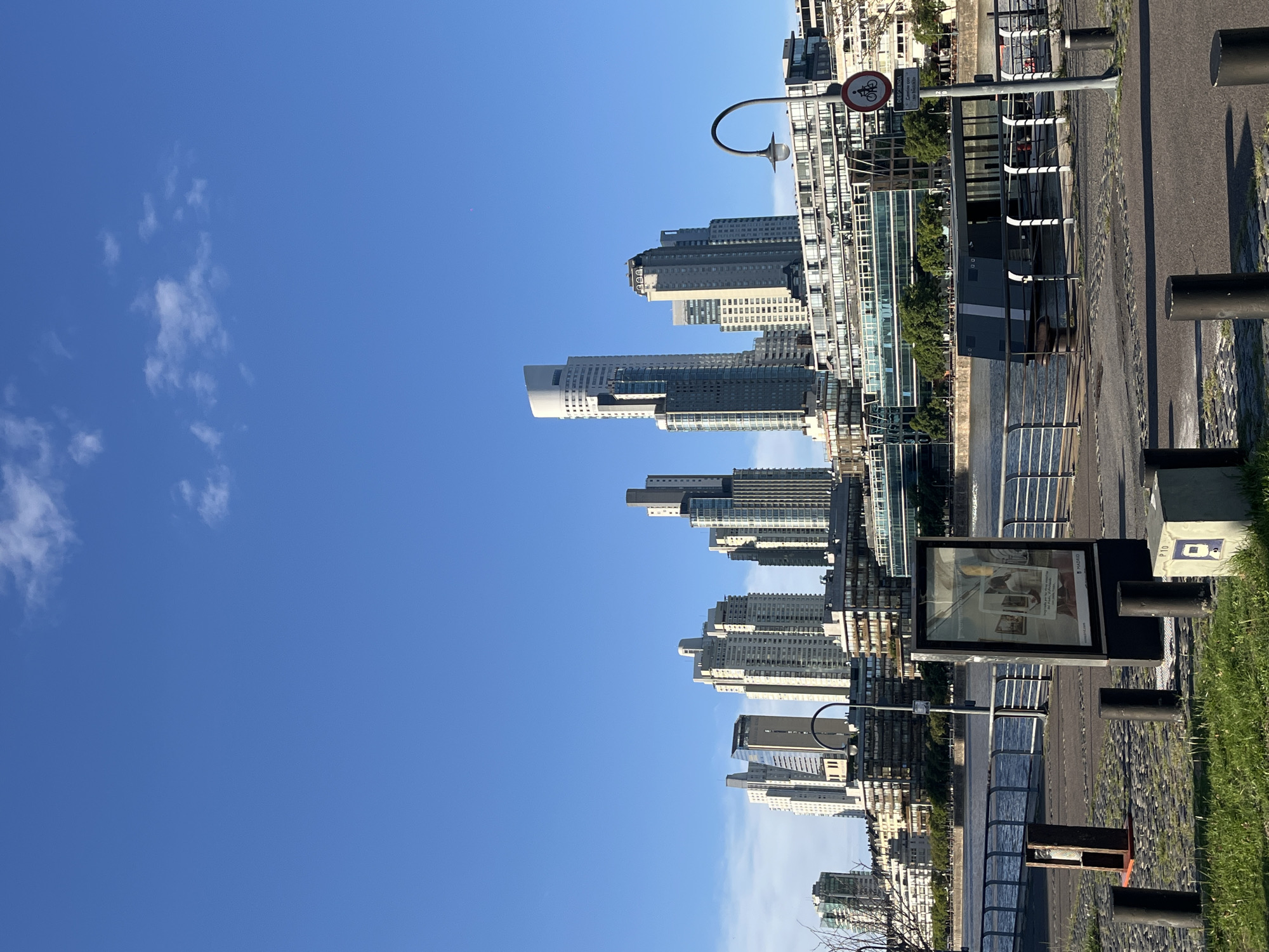 The image shows a cityscape along a waterfront in Buenos Aires, Argentina, specifically the Puerto Madero district. Modern high-rise buildings dominate the skyline. The waterfront features a pedestrian promenade with street furniture and signage. The sky is clear blue with scattered clouds. A body of water separates the promenade from the buildings.