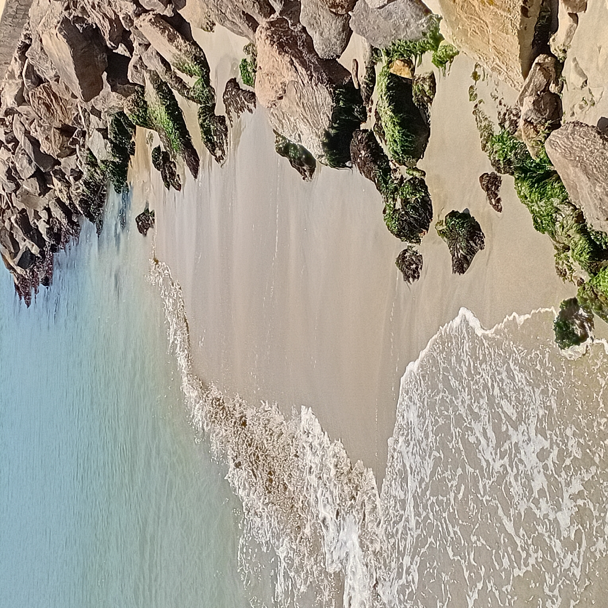 This aerial shot depicts a beach in Unknown, Unknown, where ocean waves meet the sandy shore. A rock formation lines the top of the beach, with patches of green algae growing on the rocks. The water is light blue, and the sand is light tan, with white foam from the waves creating a contrast.