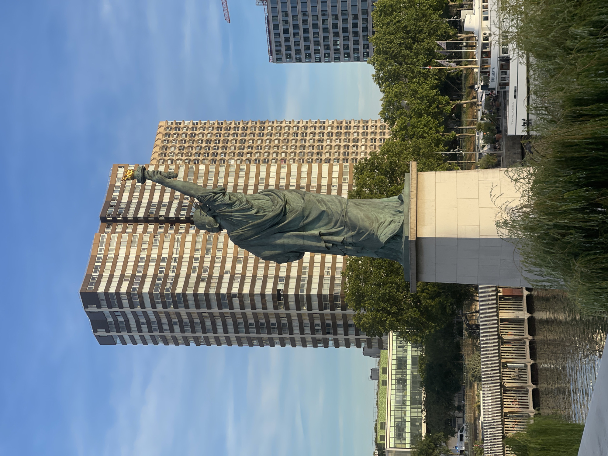 The image shows the Statue of Liberty replica on the Île aux Cygnes in Paris, France, seen from behind. The statue is a bronze sculpture with a gold torch held aloft. It stands on a stone pedestal. Behind the statue are several tall buildings, including a brown high-rise building with many windows, another brown building with a patterned facade, and a grey building under construction. Lush green trees are visible between the buildings and the river. The Seine River is in the foreground, with a white boat named "QUAI LASERTE" docked on the right. The sky is clear and blue, with a few wispy clouds. The overall scene depicts an urban landscape with a prominent landmark.