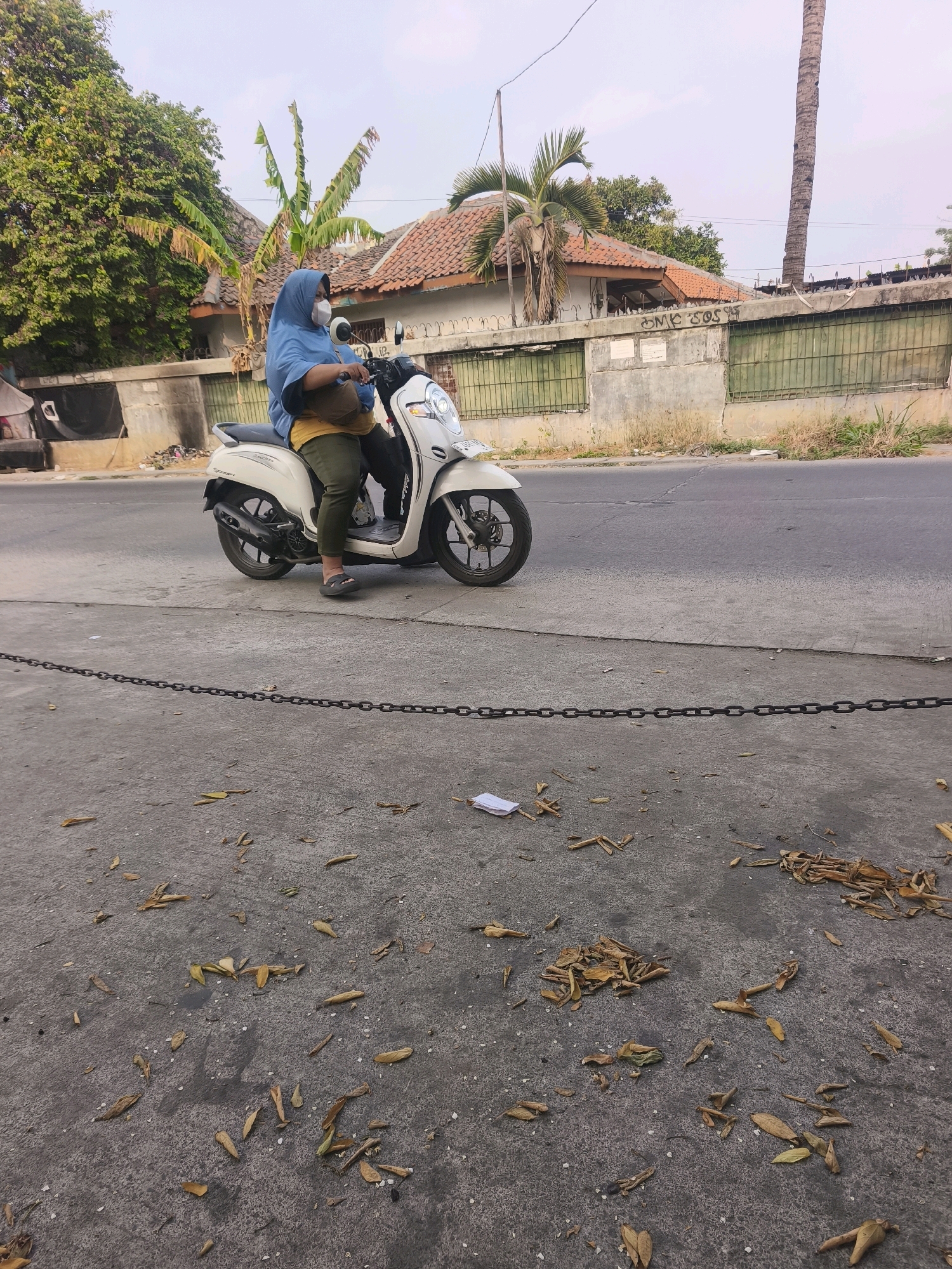 A person wearing a blue hijab and a face mask is sitting on a white scooter. They are wearing olive green pants and dark flip-flops. The scooter is parked on the side of a street in Bekasi, Indonesia. The background shows houses with tiled roofs and lush green trees, along with a tall palm tree and a concrete wall with a metal fence. The foreground is a concrete sidewalk with scattered dry leaves and a black chain lying on it. The sky is overcast.