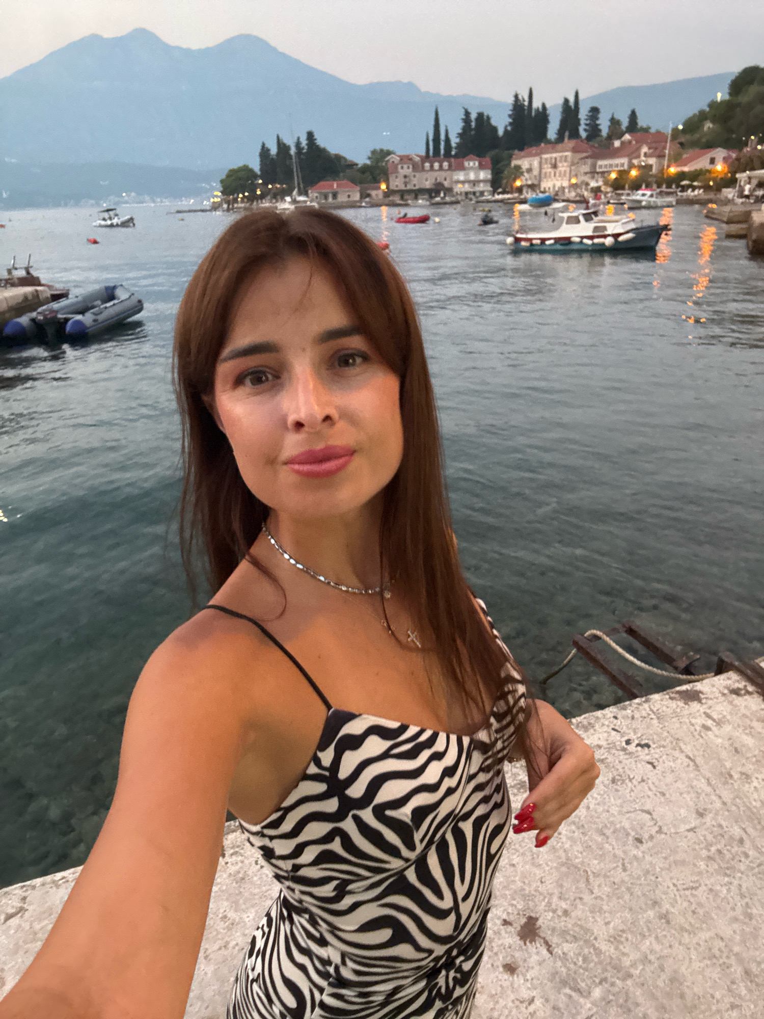 A woman with long, brown hair and a zebra-print dress is standing on a stone dock. She is taking a selfie with the water and a town in the background. The water has several boats docked on it. In the background, there are mountains and buildings. The sky is overcast. The woman has on a silver necklace with a cross charm. Her fingernails are painted red.
