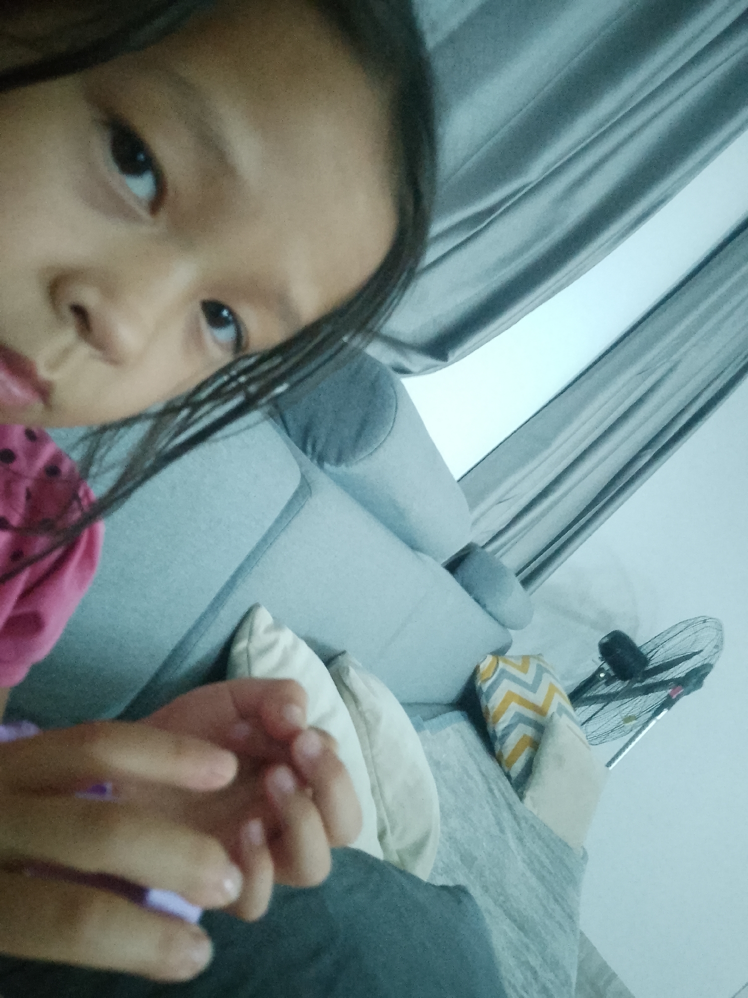 The image shows a close-up, tilted selfie of a young East Asian girl. Her face is partially visible, with her left eye and a portion of her nose and mouth in focus. She is wearing a pink shirt with black polka dots. Her hands are in the foreground, intertwined. Behind her, a light blue couch with several cushions is visible. To the right, a black oscillating fan is positioned near a window with grey curtains. The overall setting appears to be an indoor living space. There is no indication of the location being in the city of Келдемұрат, Kazakhstan.