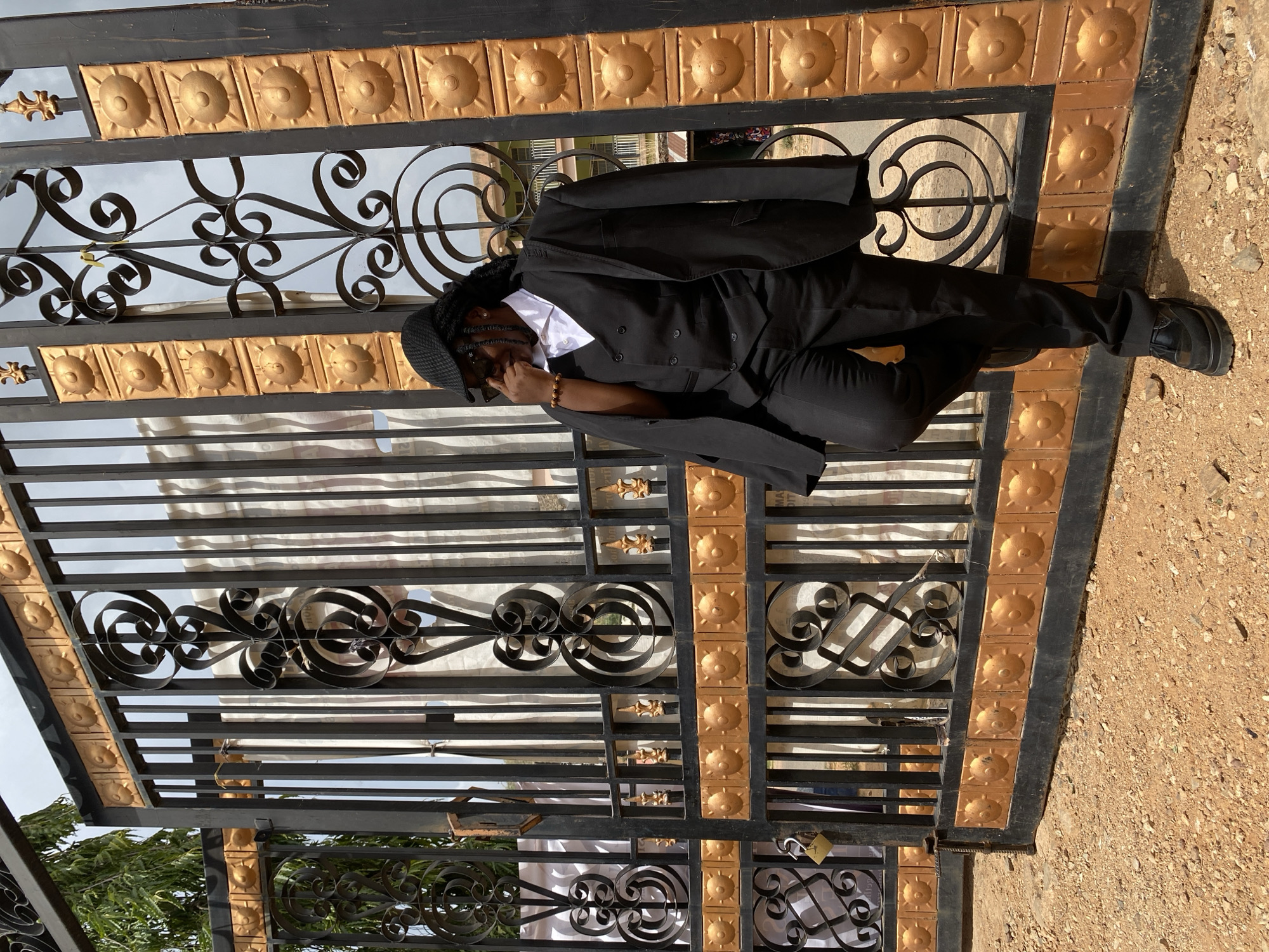 A person wearing a black suit, white shirt, black hat, and dark sunglasses stands in front of a black metal gate with ornate gold detailing. The person is leaning against the gate with their right leg extended, left leg bent. The gate has a padlock and intricate ironwork. The ground adjacent to the gate is covered in reddish-brown gravel. Vegetation is visible in the lower-left corner. The scene is located in the city of Gwong, Nigeria.