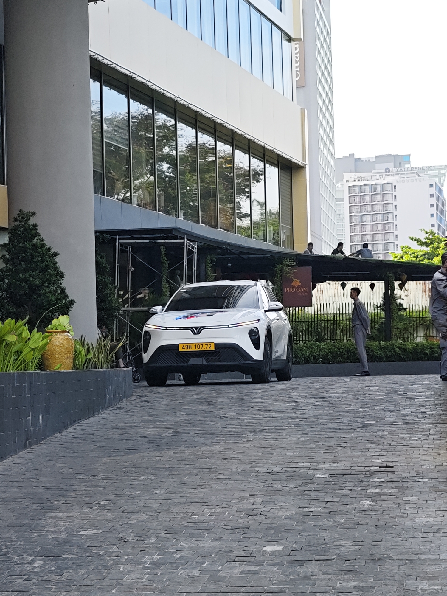 The image depicts an outdoor scene in Nha Trang Ward, Vietnam, likely in front of a modern building with large glass windows and a concrete facade. A white electric vehicle, possibly a VinFast, with a yellow license plate reading "49H-107.72" is parked on a textured grey paved driveway. A man in a grey uniform stands to the right of the vehicle, facing away from the camera. Above the vehicle, a covered outdoor area with seating is visible, where three individuals are seated. A sign with Vietnamese text, "PHO GAM," hangs from the covered structure. The environment includes potted plants and a stone wall bordering the driveway. In the background, tall buildings are visible, including one with "HYATT REGENCY" and "NOVOTEL" signage. The sky is bright and appears to be daytime.