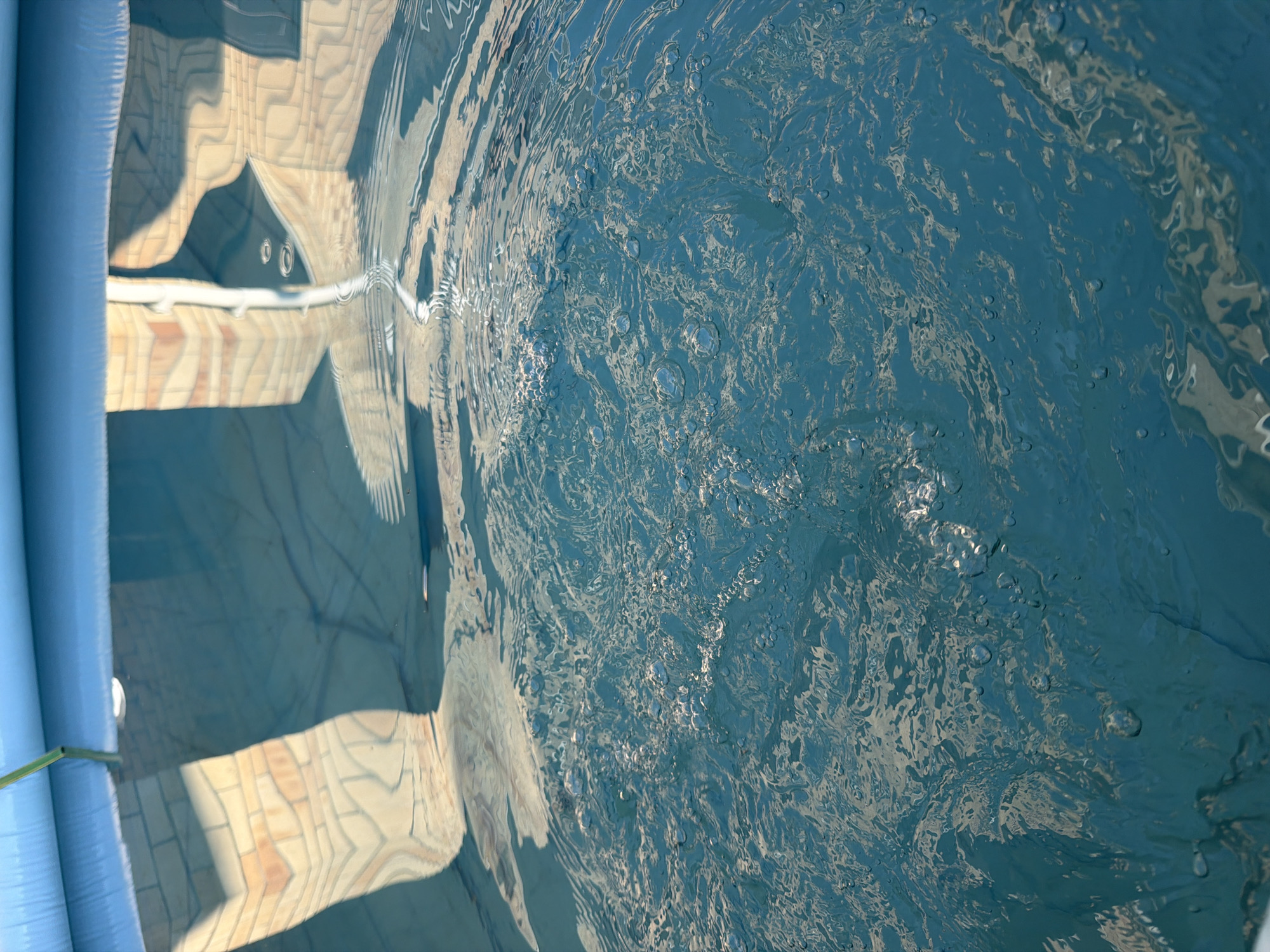 The media file shows an abstract close-up of rippling blue water in an above-ground swimming pool. The water is disturbed, with many small bubbles visible near the surface, suggesting recent activity. The lighting indicates it is daytime, with bright sunlight reflecting off the water, creating shimmering patterns and distortions.
To the left of the image, a portion of the pool's blue, inflatable edge is visible. Reflected on the surface of the water, and distorted by the ripples, is a pattern that resembles tiled flooring or a brick pattern, likely from the surrounding environment. There are also indistinct shadowy shapes reflected, possibly from nearby structures or objects. The overall impression is one of a bright, sunny day, with the focus on the dynamic and textured surface of the water.
There is no visible text in the image. The scene is outdoors, likely in a residential or recreational setting. The activity suggested is swimming or some form of water play that would cause the water to be disturbed and bubbly.