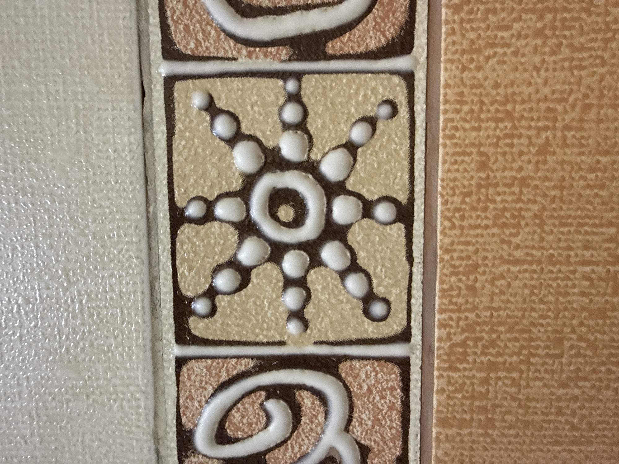 The image displays a close-up view of decorative tiles, likely from a building interior. The primary focus is on a central tile featuring a white, raised pattern resembling a stylized sun or starburst on a light beige background. This pattern is outlined in dark brown. To the left and right of this central tile are adjacent tiles with a textured, light brown and reddish-brown speckled pattern, also framed by dark brown and white borders. Above this row of tiles is a textured, off-white tile. Below, a solid, textured light orange tile is visible. The scene appears to be a static, architectural detail. There are no people or actions depicted. The lighting is even, suggesting an indoor environment. The location is Taldybulak, Kazakhstan, but the specific context within the city is not discernible from the image alone.