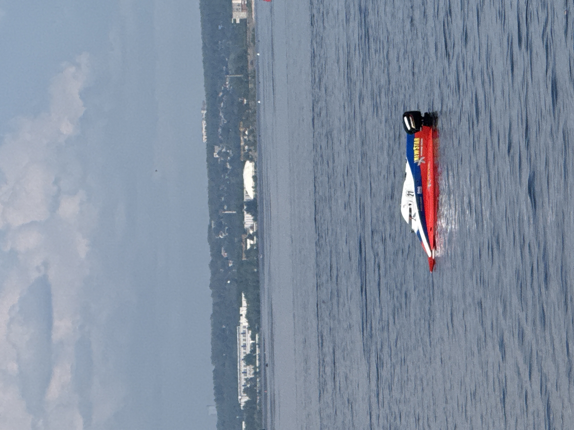 A white and red speedboat with the number "21" and "NEW STAR" printed on its side is positioned in the middle of a body of water. The boat has a black outboard motor. In the background, a tree-lined shore is visible with buildings and structures. The sky above is cloudy. The scene appears to be a body of water, possibly the Kama River, in the city of Perm, Russia.