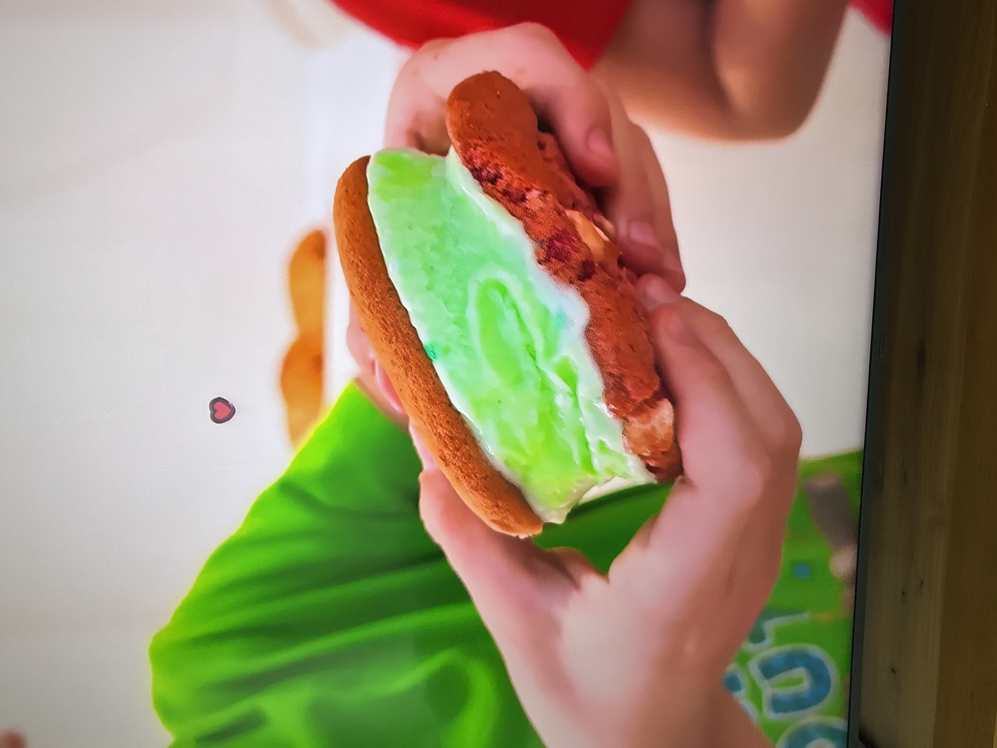 The image shows a close-up of a person's hands holding a green ice cream sandwich. The sandwich consists of two dark brown cookies with a thick layer of bright green ice cream in between. The ice cream is starting to melt and drip down the sides. In the background, another person is visible wearing a red shirt, and part of a person in a green shirt can be seen. There is a blurred white background with a small pink heart graphic overlaid. The scene appears to be indoors, possibly in a kitchen or a studio setting, given the controlled lighting and plain backdrop. There is no indication of the image being taken in Dubai, United Arab Emirates.
