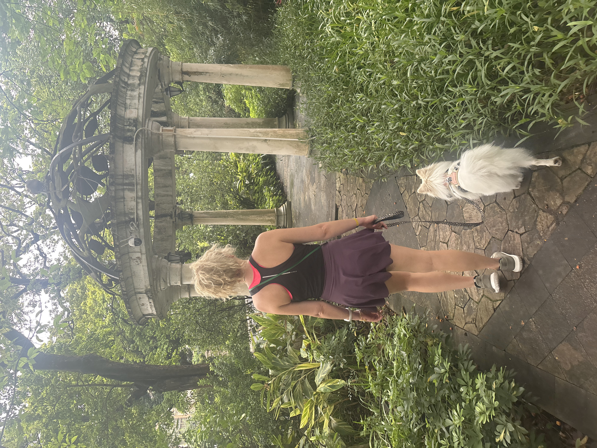 A woman with blonde, curly hair pulled into a ponytail is walking away from the camera on a stone path in a lush, green garden. She is wearing a black athletic tank top over a pink sports bra, a purple skirt, and grey athletic shoes. A white Pomeranian dog with a pink harness is walking beside her on a black leash. To the right of the path is dense, tall green foliage, and to the left are tropical-looking plants. In the background, a weathered, ornate stone gazebo with columns and a domed roof stands amongst the trees. The sky is overcast, suggesting a cloudy day. The scene appears to be outdoors, possibly in a park or botanical garden, and is devoid of any visible signage or context that would place it specifically in Nanhai District, China.