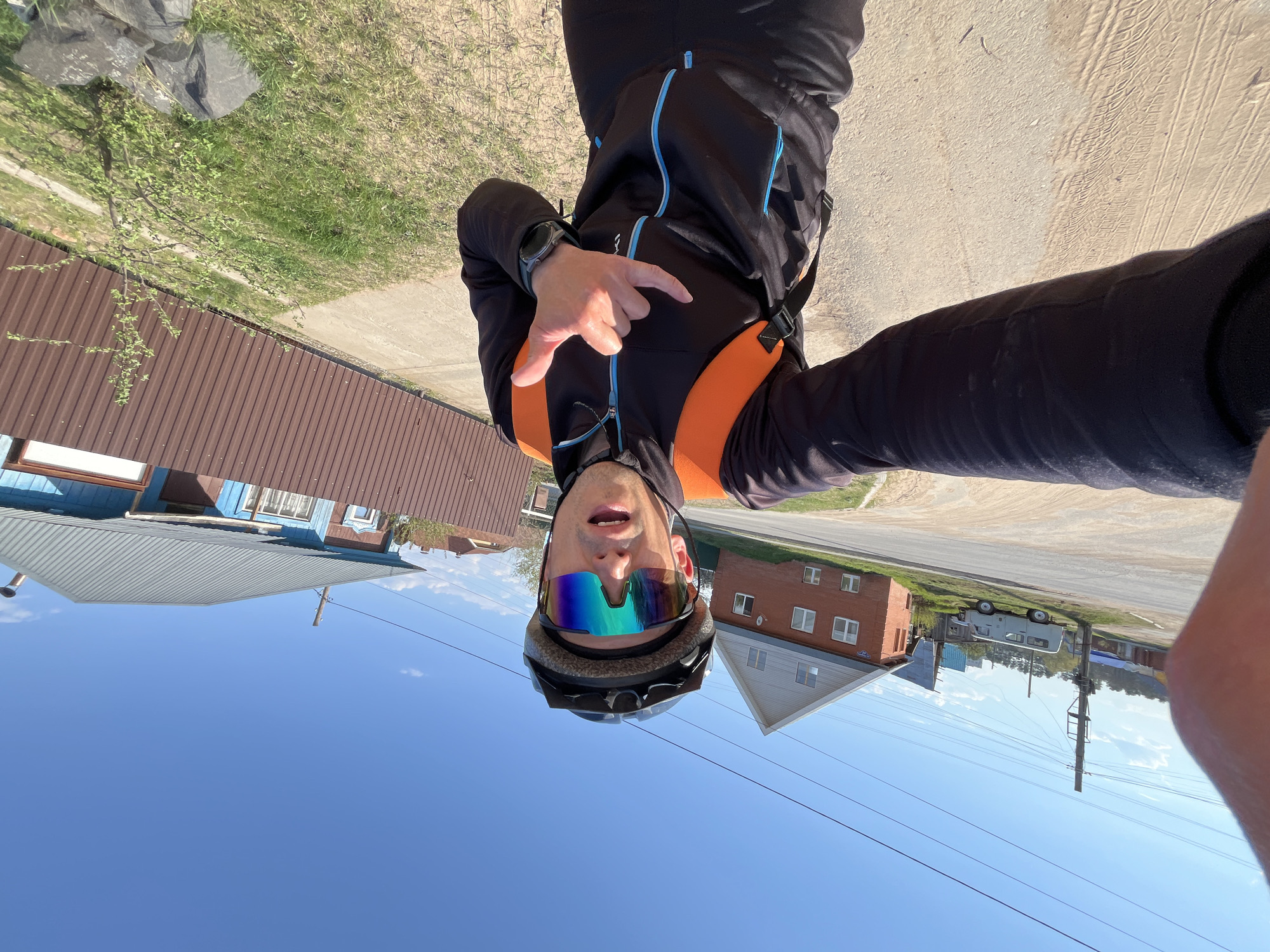 A man in cycling gear, including a helmet and reflective sunglasses, is taking a selfie outdoors in what appears to be the city of Chaikovsky, Russia. He is wearing a black jacket with orange and blue accents. He is making a hand gesture. The background shows a mix of residential buildings with various roof types, green grassy areas, a dirt or paved road, and a clear blue sky. Power lines and utility poles are visible in the distance. Based on the architecture and layout, this could be a suburban or semi-rural area within Chaikovsky.