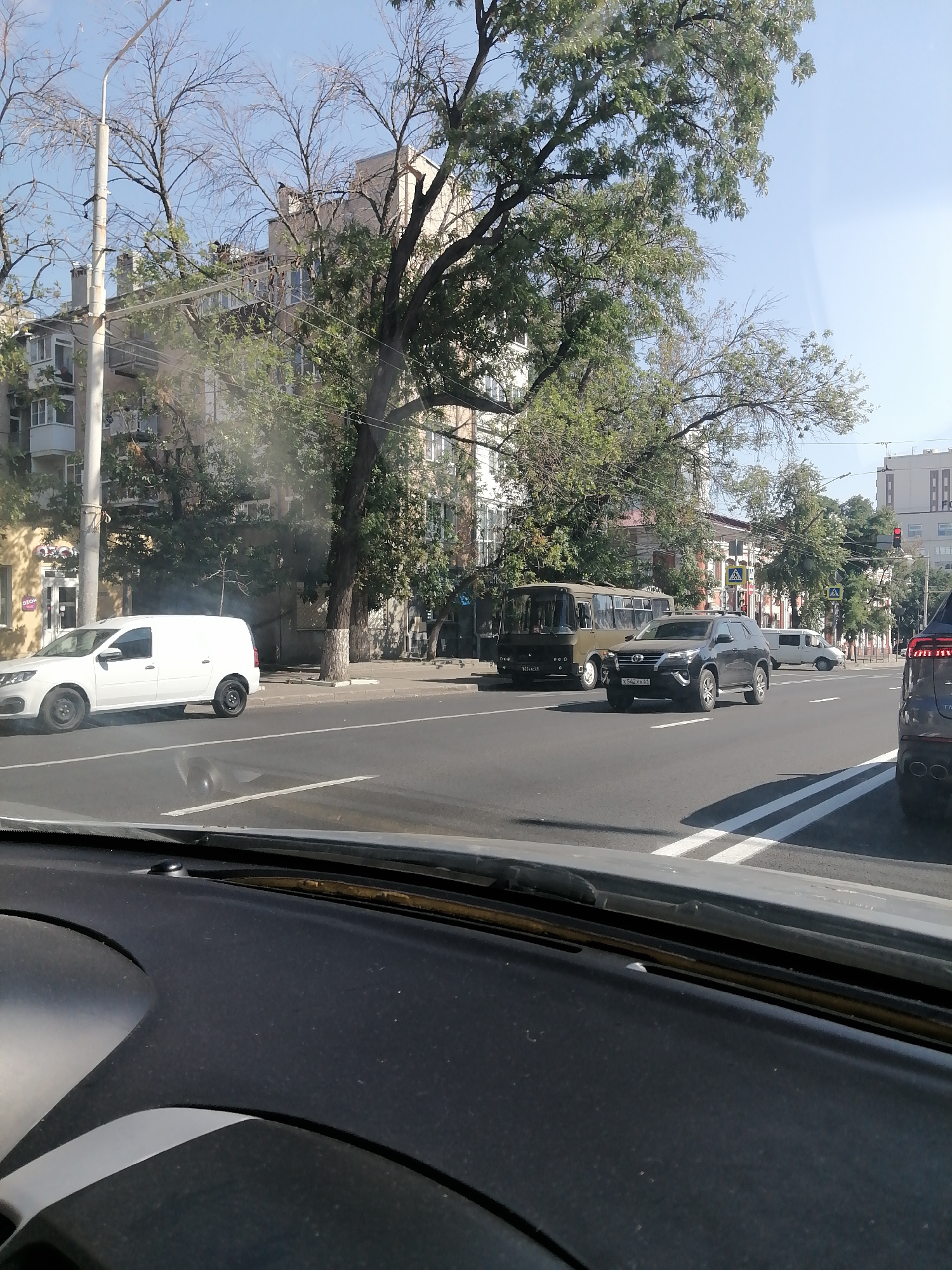 The image shows a street scene in Rostov-on-Don, Russia. A white cargo van is parked on the side of the road, with smoke emanating from its rear. In the background, a green bus and a dark SUV are visible, along with several multi-story buildings and trees lining the street. The sky is clear and blue. The perspective suggests the photo was taken from inside another vehicle.