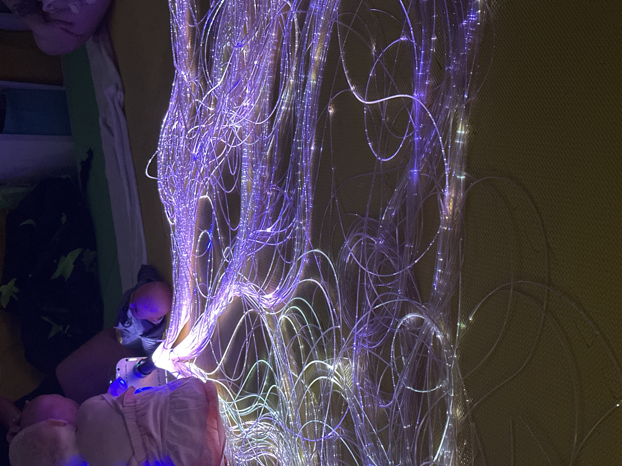 The image shows a close-up view of a collection of fiber optic lights, predominantly illuminated in shades of purple and white. These lights are arranged in a dense, tangled mass, creating a visually dynamic and somewhat abstract composition. The lights appear to be on a textured surface, possibly a mat or rug, which has a yellowish-brown hue. In the background, partially visible, are a few figures, including what appears to be a child in a pink dress, seated on the floor. There are also some dark fabric items and possibly other soft furnishings in the background. The lighting is low, accentuating the glowing nature of the fiber optics. There is no discernible location in the city of Wakefield, United Kingdom mentioned or indicated within the image.