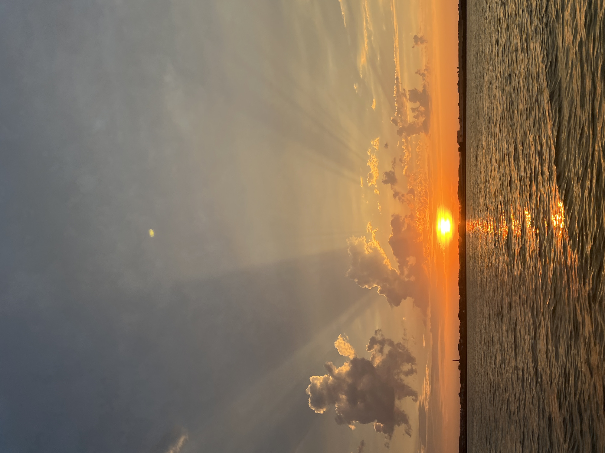 A serene sunset over the waters near Novopichugovo, Russia, paints the sky with a gradient of blues, oranges, and yellows. Sunbeams pierce through scattered clouds, casting dramatic rays across the scene. The sun, a bright orb, is low on the horizon, its reflection shimmering on the rippling surface of the water. A distant silhouette of buildings and structures forms a faint line between the water and the sky, hinting at the presence of a settlement. The overall impression is one of tranquility and natural beauty.