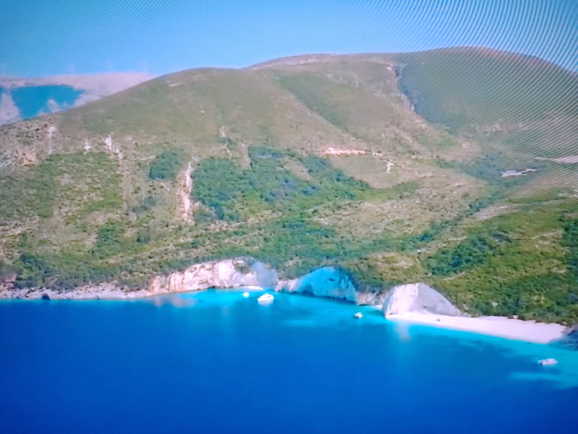 An aerial view captures a rugged, green-covered mountain meeting a vibrant blue sea. White cliffs and a sandy beach are nestled along the coastline, with a few small boats dotting the water. The sky is a clear, pale blue. This scene depicts a coastal landscape, however, the provided context of the city of Huntingdonshire, United Kingdom is inaccurate as Huntingdonshire is an inland county and does not have a coastline.