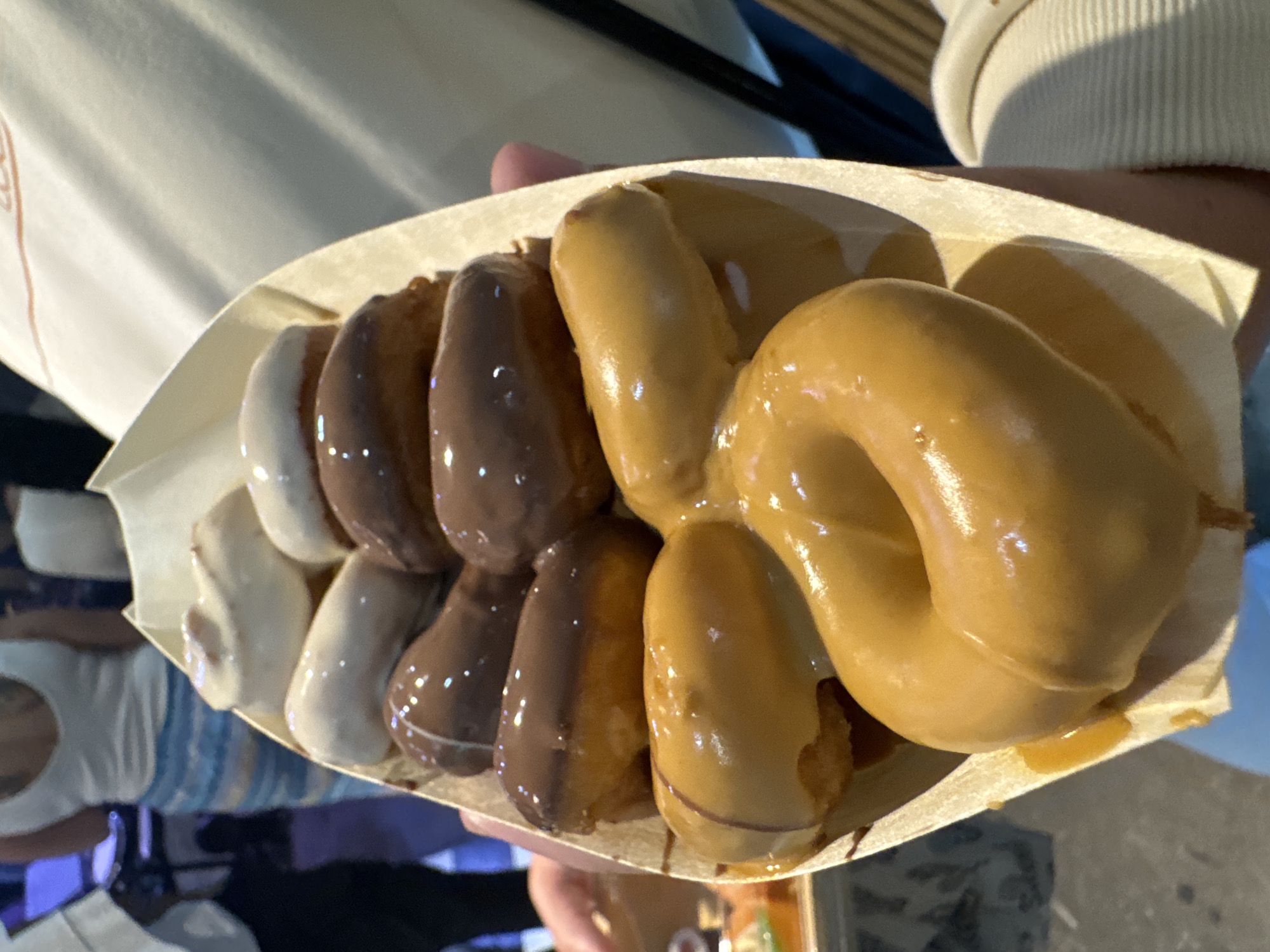 A close-up image shows a person holding a biodegradable paper tray filled with a variety of glazed donuts or churros. The person's arms are partially visible, wearing light-colored clothing, including what appears to be a ribbed sweater on the right side.
The tray contains numerous elongated, bite-sized desserts, arranged in rows. A significant portion of the desserts, including a large, prominent ring-shaped donut, are covered in a thick, shiny, light brown or caramel-colored glaze that appears slightly gooey. Other desserts in the tray are coated in a white glaze and a dark chocolate glaze, creating a diverse assortment of flavors. The glazes reflect light, giving them a glossy appearance.
The background is dark and blurred, suggesting an indoor setting or an evening/nighttime outdoor event, possibly a market or a dessert shop. The artificial lighting is evident from the reflections on the glazes. The image focuses entirely on the prepared desserts. No text is visible in the frame. The scene is likely set in Cape Town, South Africa.
