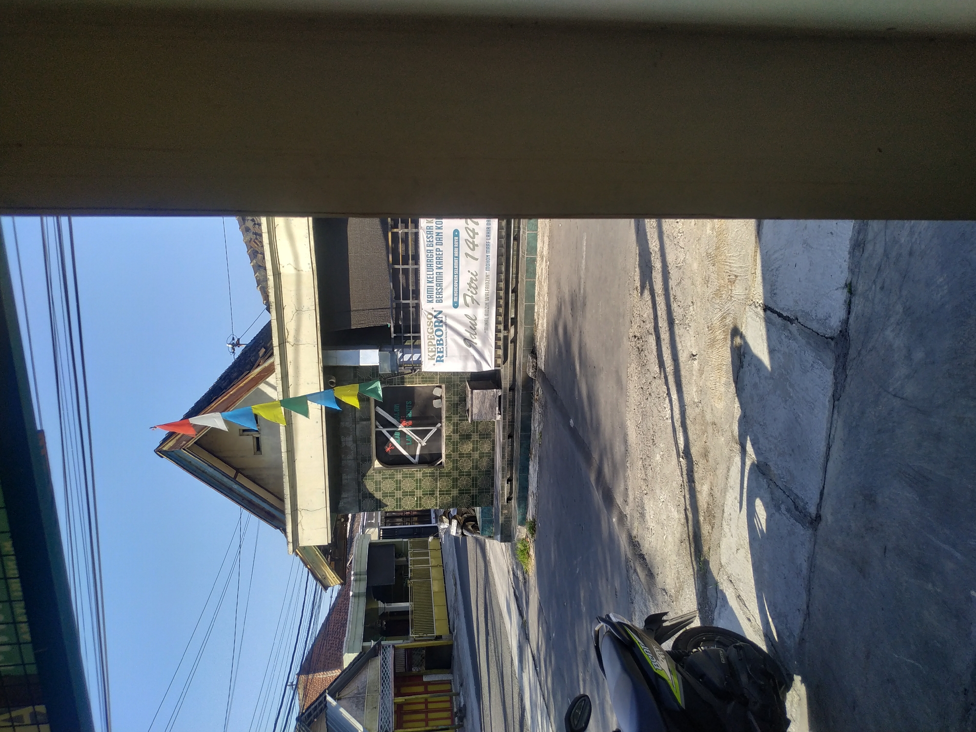 A bright, sunny day in Batan, Indonesia, is captured in this image, which depicts a street scene with a few buildings and a parked motorcycle. The foreground features a portion of a building's wall on the right, obscuring part of the view. To the left, a black and green motorcycle is parked on the sidewalk, its front tire and handlebars visible. The street is paved and shows signs of wear, with shadows cast by the motorcycle and trees.
In the middle ground, a building with green tiled walls stands out. It has a prominent roofline and a large banner hanging on its facade. The banner displays text in Indonesian, including "Idul Fitri 1447," indicating a celebration of Eid al-Fitr. Small triangular flags in red, white, blue, and yellow are strung across the top of the building. Another smaller building with a red-tiled roof is visible behind it, with houses and fences further in the background. Power lines crisscross the clear blue sky, a common sight in urban Indonesian landscapes.