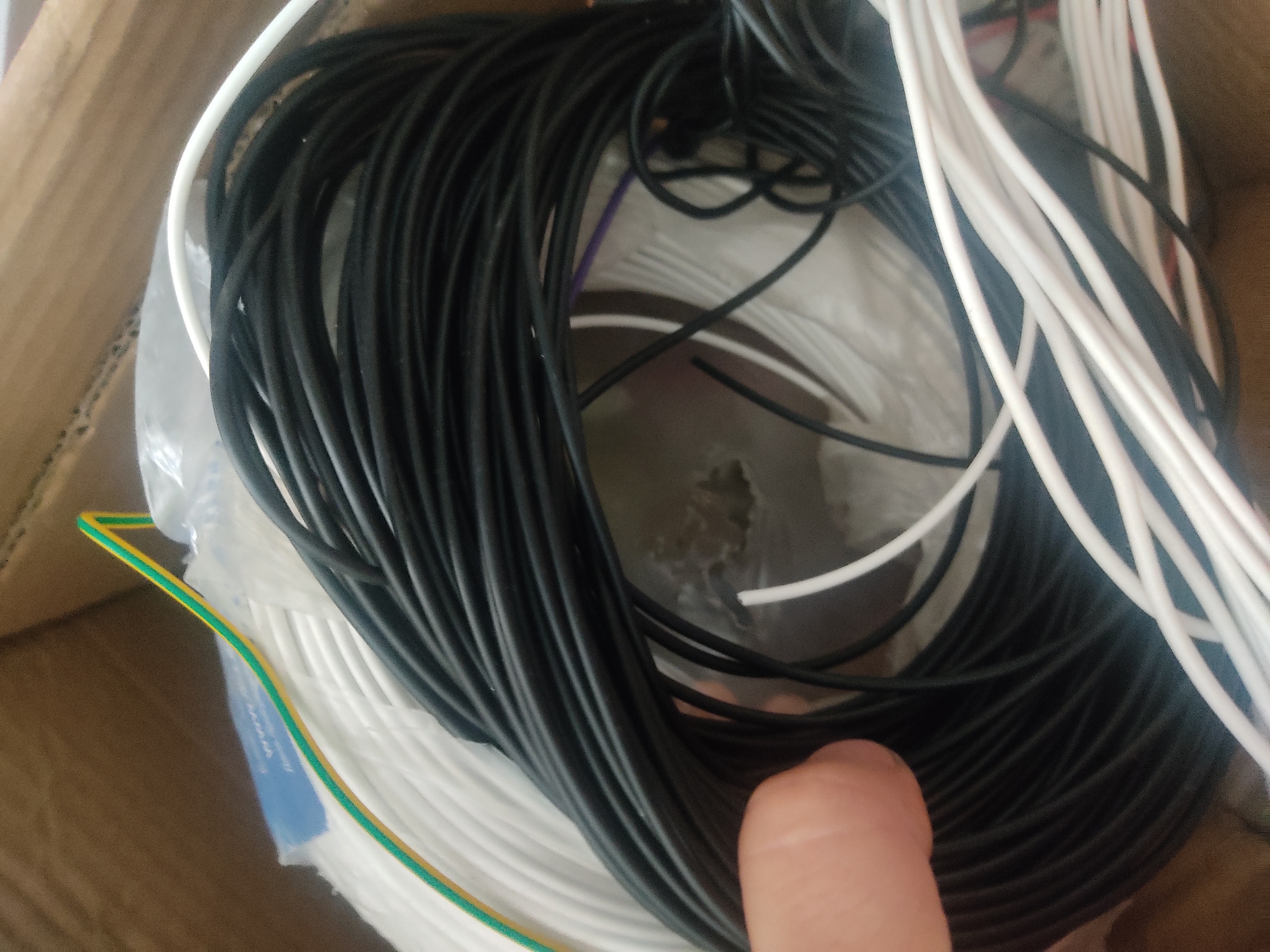 The image shows a close-up, overhead view of a collection of wires, primarily black and white, coiled together within a cardboard box. A human finger is visible in the lower right corner, partially obscuring some of the wires. The wires appear to be insulated and are neatly arranged in coils, suggesting they are intended for electrical or electronic use. There is also a section of green and yellow striped wire visible along the left edge, which is often used as grounding wire. The setting appears to be indoors, given the lighting and the presence of a cardboard box, but the specific location is not identifiable. No text is visible in the image. The activity taking place is unclear, but the organized nature of the wires implies they are either being stored, prepared for use, or have been recently organized. The overall impression is one of a practical, functional scene related to electronics or electrical work.