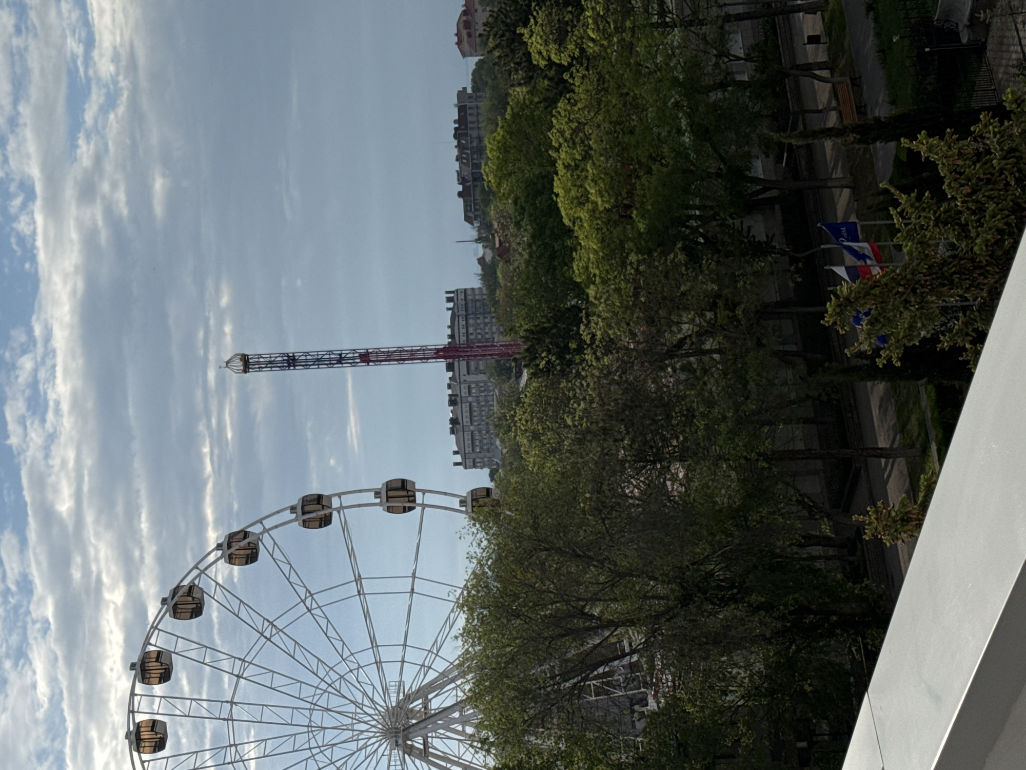 The image shows a view from what appears to be a high vantage point, possibly a building or elevated structure, in Yevpatoriya, Ukraine. A large Ferris wheel is prominent in the foreground, with its support structure visible behind some trees. A tall drop tower ride is also visible in the background, with red and blue coloring. Trees and building line the background of the scene, with the sky above filled with clouds. Flags stand to the right of the image behind the trees.