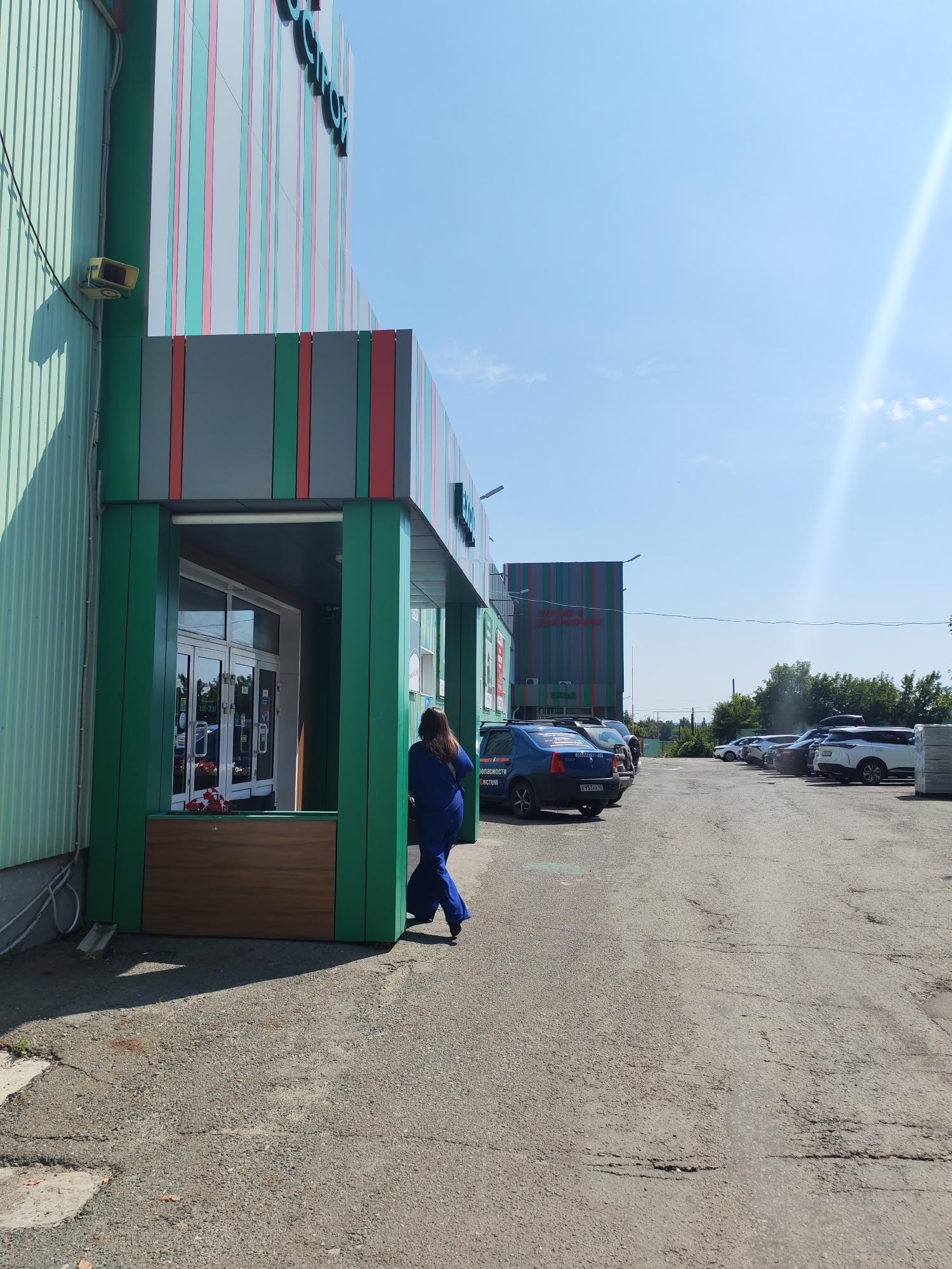A medium shot captures a woman in a blue outfit walking away from the entrance of a building with a green facade on a sunny day. The building has colorful vertical stripes of red, pink, and gray on its upper section. The woman is walking on an asphalt surface that shows signs of wear and tear, with cracks and patches visible. A parking lot is to the right of the building, with several cars parked. The sky is clear and blue with a bright sun causing lens flare. In the background, trees are visible, suggesting a location with some greenery. The overall scene is of an outdoor commercial or retail environment.