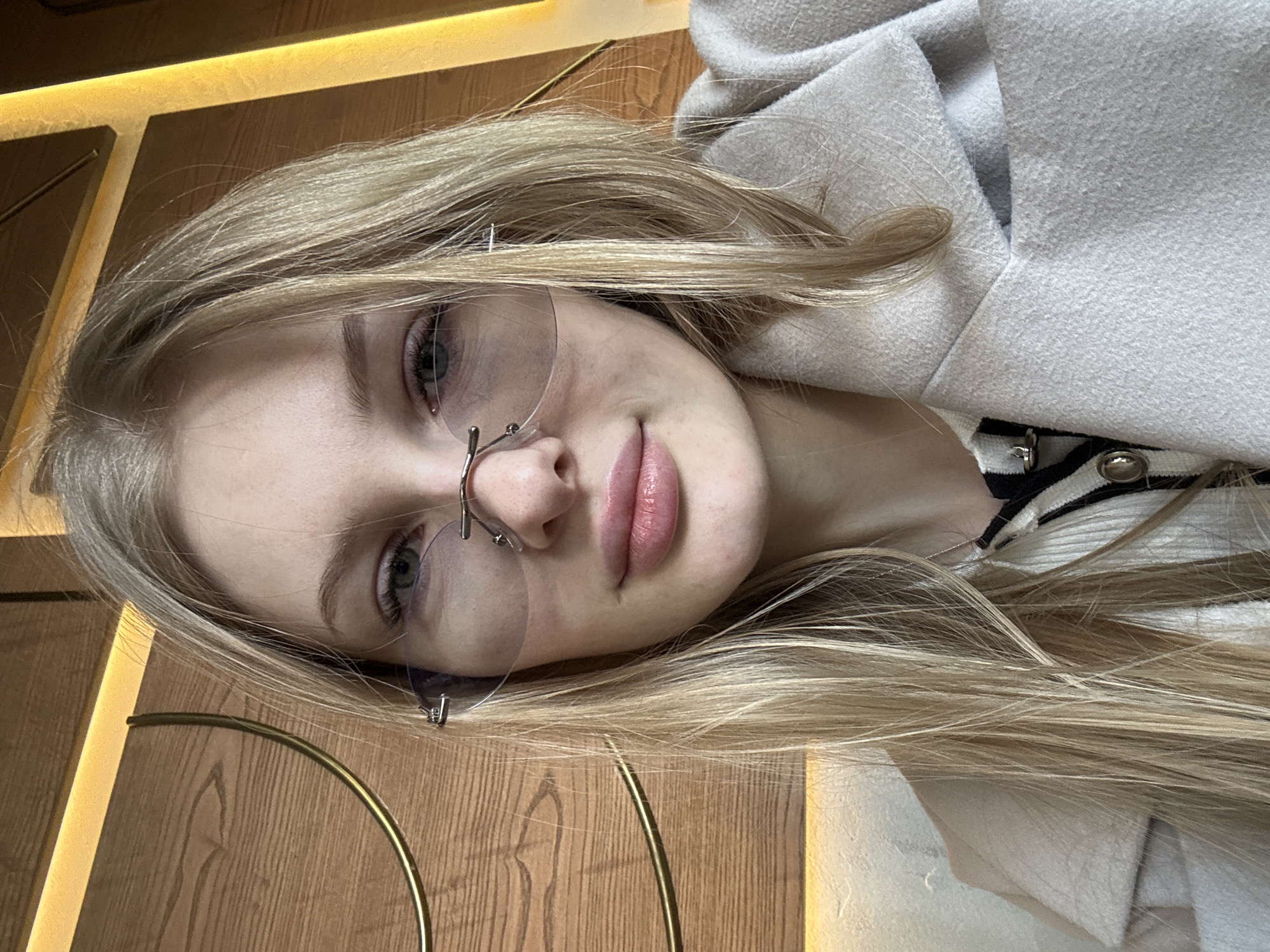 A close-up photograph shows a young woman with light blonde hair and pale skin. She is wearing frameless eyeglasses with nose pads. She is looking directly at the camera with a neutral expression. The background includes a wooden panel with gold trim. She appears to be wearing a white coat or sweater with a black and white striped undergarment visible. There are no other people, subjects, objects, actions, interactions, or environmental elements apparent. There is no information in the image that identifies the location as Perm, Russia.
