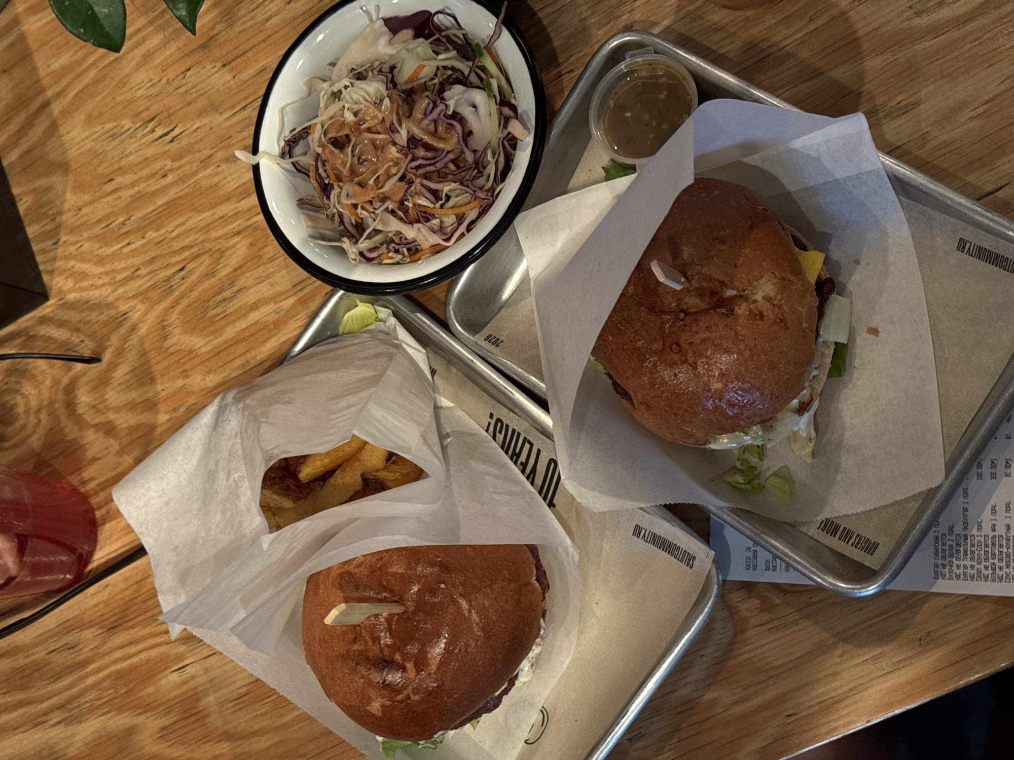 The image depicts a table setting in what appears to be a restaurant. A wooden table serves as the backdrop, with two burgers, a side of French fries, a small bowl of salad, and a dipping sauce container arranged on metal trays. The burgers are served in white paper bags, with wooden skewers holding them together. The salad consists of shredded cabbage and other vegetables, topped with dressing, and is served in a white bowl with a black rim. A partially visible receipt lies near one of the trays, containing Cyrillic text. The receipt indicates that the location might be Nizhny Novgorod, Russia, due to the mention of "BURGERS AND MORE" and Cyrillic script. A glass with a red liquid is partly visible on the left side of the image. A plant is also seen in the upper left corner.