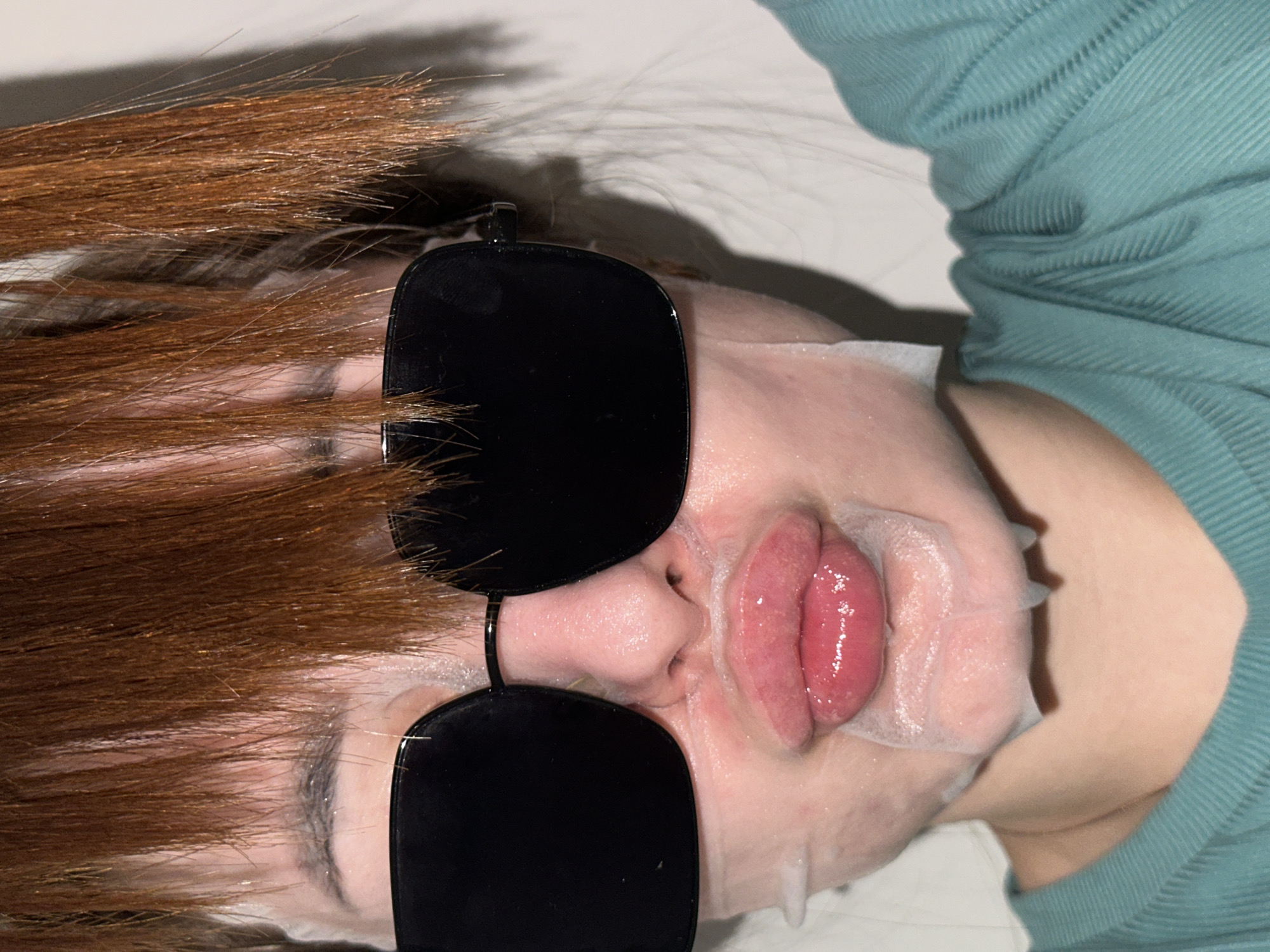 An upward-facing, close-up shot of a person lying down, engaged in a self-care routine in Chaikovsky, Russia. The person is wearing a white sheet face mask, which covers most of their face except for their nose and mouth. They have large, black, opaque sunglasses placed over the mask, completely obscuring their eyes.
The person's long, reddish-brown hair is spread out, framing their head. Their lips are slightly parted, revealing a shiny, pinkish-red color, and some serum from the face mask is visible around the mouth and jawline. They are wearing a teal or light green ribbed top, part of which is visible on the right side of the frame. The overall impression is one of relaxation or a beauty treatment. The background appears to be a light, plain surface, possibly a bed or pillow. No text is visible in the image.