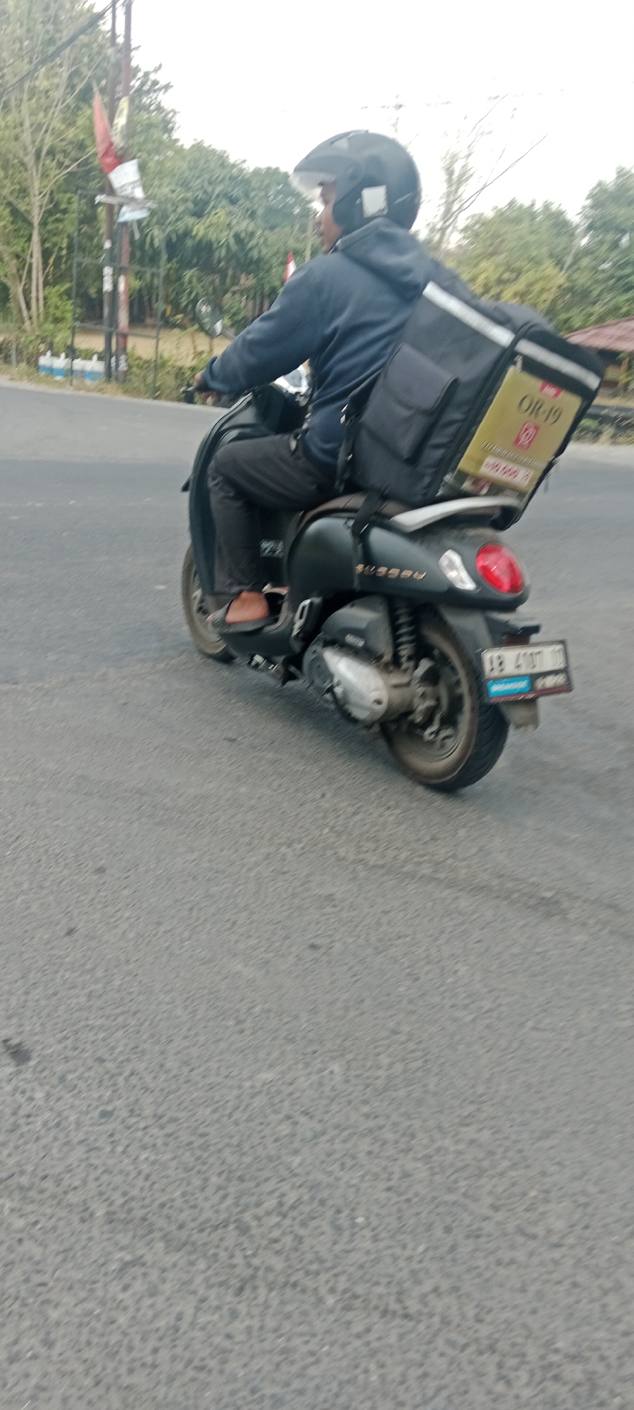 A person wearing a blue hooded jacket and a black helmet rides a dark-colored scooter on a paved road. The scooter has a large black cargo box mounted on the back, with a yellow panel on its side displaying text and numbers, including "OR-19". The scooter also has a license plate that reads "AB 4187 BI". The background shows trees and a cloudy sky, suggesting an outdoor environment in Maguwoharjo, Indonesia. The road appears to be asphalt.