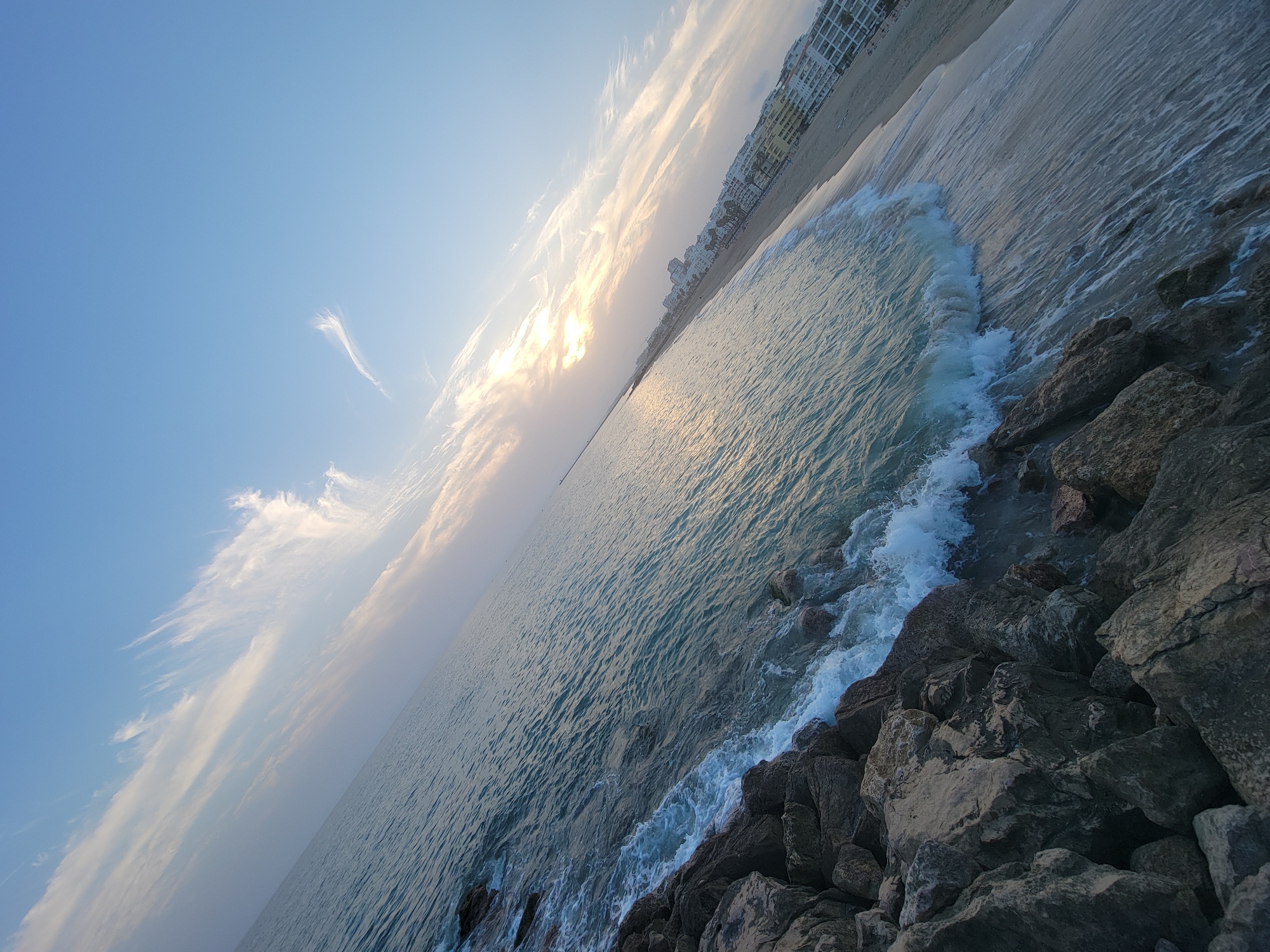 An eye-level, wide shot captures a diagonal view of the sea meeting a rocky shoreline, with a beach and buildings in the distance under a hazy, pale blue sky with wispy white clouds. The sun is low in the sky, casting a warm glow on the scene and reflecting on the water. Waves gently crash against the rocks, creating white foam. The buildings appear to be a resort or apartment complex, consistent with the coastal landscape of Quarteira, Portugal. There are no people visible in the image. The overall mood is serene and tranquil.