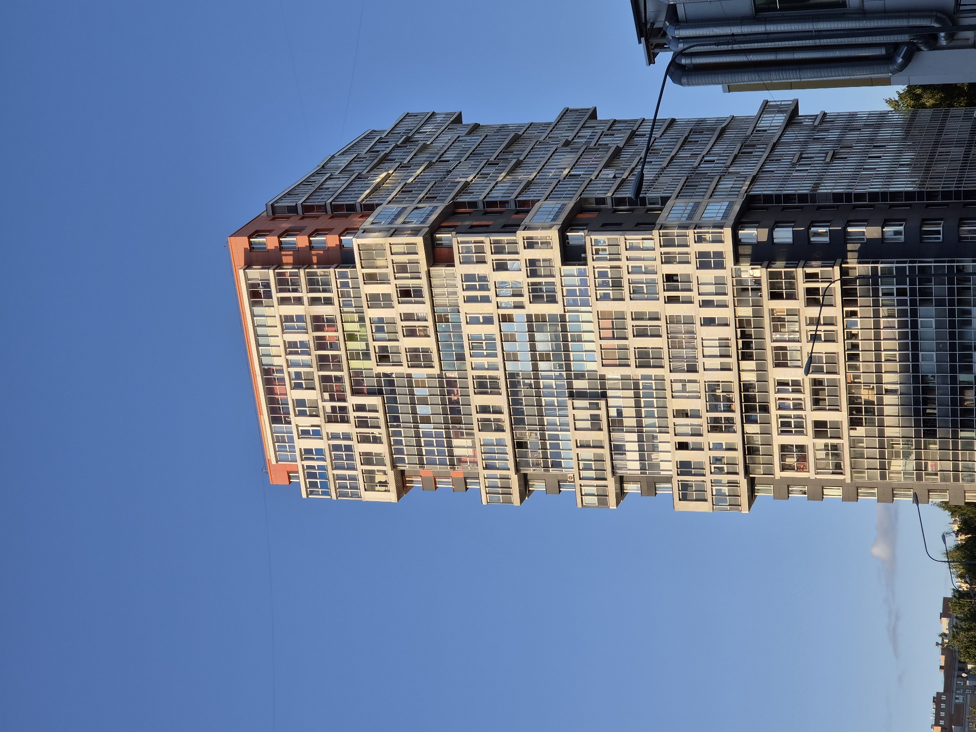 A tall, modern apartment building dominates the frame against a clear blue sky in Kudrovo, Russia. The building features a distinctive facade with a staggered, geometric design, characterized by numerous windows and balconies. The upper floors are accented with orange cladding. To the right of the apartment building, a portion of another structure with large, corrugated pipes is visible. The lower section of the image shows the tops of trees and hints of other buildings. A street lamp with a curved arm extends across the lower right portion of the scene. The lighting suggests it is late afternoon or early evening, with shadows cast on the building. No people are visible.