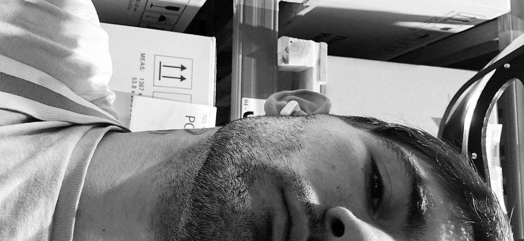 This black and white photograph, taken in Plovdiv, Bulgaria, presents a close-up, sideways view of a man's head and upper torso. The man's face is partially visible, showing his right eye, part of his nose, and a stubbly beard or close-cropped hair on his jawline. His dark hair appears slightly damp or styled. A white earbud or hearing aid is prominent in his left ear. He is wearing a light-colored, short-sleeved shirt with subtle horizontal stripes.
The man appears to be resting or lying down, with his head tilted approximately 90 degrees to the right. The background features stacked cardboard boxes, indicating an indoor setting, possibly a storage area, workshop, or a room with packed items. One white box clearly displays shipping details including "MEAS: 1367 X 53.8 X" and a "this way up" symbol with two vertical arrows. Other visible text on the boxes includes "PC" and "705-", as well as "H4-" on a label near the man's ear. To the far right, a curved, dark object with what looks like metallic studs or bolts is partially visible. The lighting suggests an indoor ambient light source, casting subtle shadows.