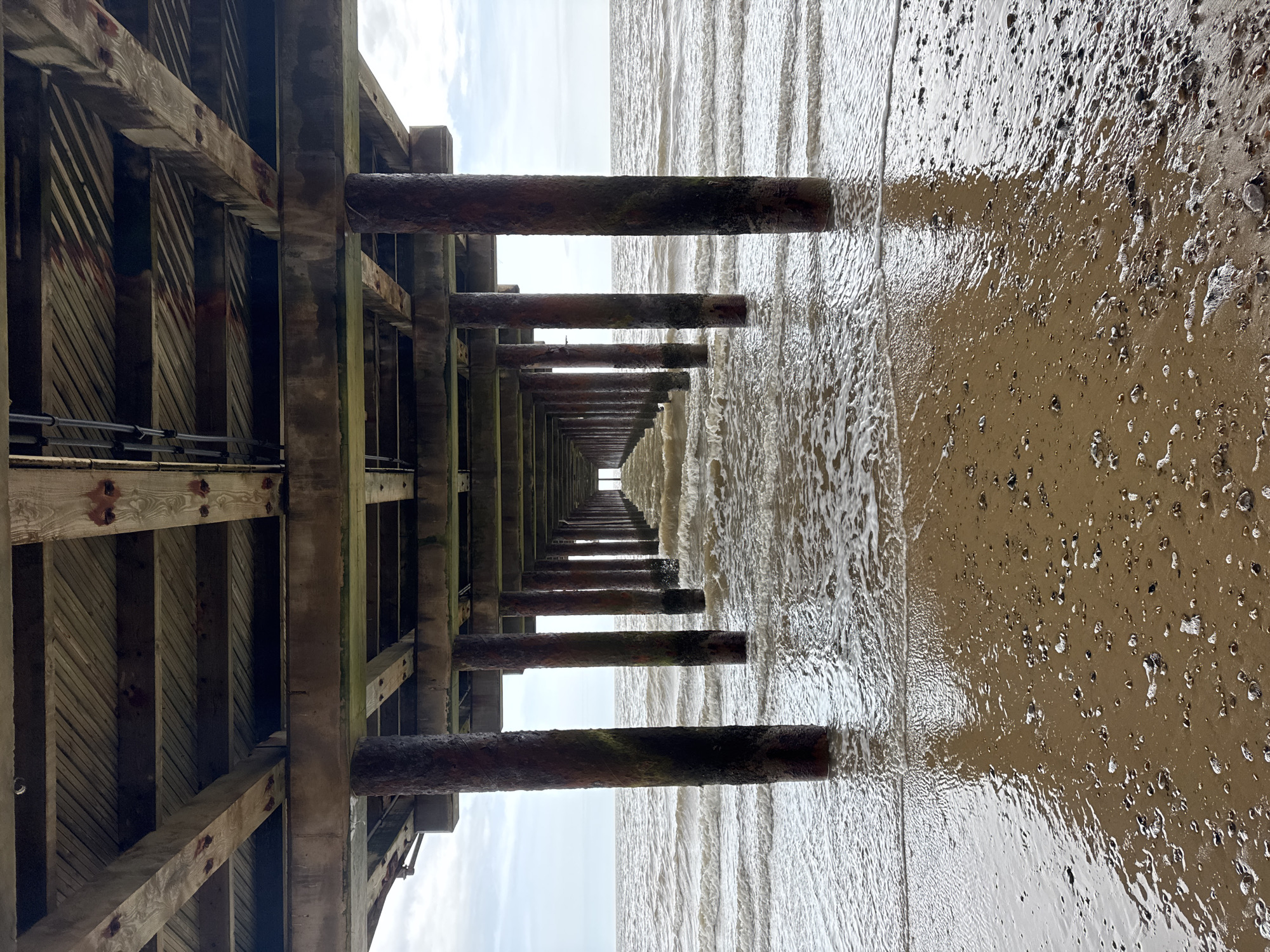 This media file captures a low-angle perspective from underneath a pier, looking out towards the sea and horizon. The scene is set during daytime, likely overcast, at a coastal location in East Suffolk, United Kingdom.
The prominent subject is the pier's understructure, consisting of weathered wooden beams, vertical supports, and diagonal bracing. Some elements appear to be concrete or stained by rust from metal fixings. This framework creates a repetitive, linear pattern that recedes into the distance, guiding the viewer's eye.
Numerous cylindrical pillars, heavily corroded and stained by rust or marine growth, extend vertically from the pier into the water and wet beach. These piles also diminish in size towards a distant vanishing point, enhancing the depth and perspective of the view.
To the right of the image, the sea meets a sandy or muddy beach that is wet and reflective, scattered with small shells or stones. Gentle, murky, brownish-grey waves break on the shore, creating white foam. The distant sky is bright and appears overcast, providing soft, diffused light.
No people or explicit activities are depicted, focusing instead on the architectural structure and the natural coastal environment. There is no visible text. The overall color palette is dominated by earthy tones of grey, brown, and dull green from the weathered wood, rusted metal, and wet sand, contrasted with the lighter grey of the sky and white of the sea foam.