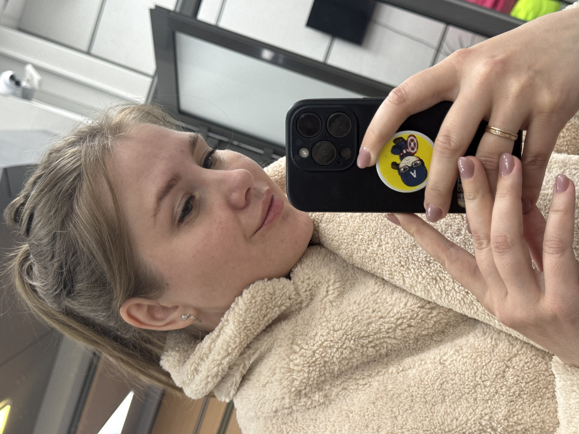 A young woman is captured in a mirror selfie, holding a black smartphone with her right hand. Her light brown hair is styled back, and she has a gentle, serene expression, looking towards the phone screen. She wears a cream-colored, fluffy sherpa-style jacket or hoodie.
Her right hand, holding the phone, displays neat, light pink manicured nails and a thin gold-colored ring with small embedded stones on her ring finger. Her left hand is also visible, resting near the phone. The phone case features a distinctive yellow circular sticker or PopSocket depicting a cartoon-style character resembling Captain America, with a blue mask and shield. The phone itself appears to be a model with a prominent triple-camera array on the back.
In her left ear, a small, silver-colored hoop earring with a tiny heart charm is visible. The background, reflected in the mirror, shows indoor elements such as white and gray walls or shelving, along with dark, rectangular frames that suggest a commercial or public indoor setting, possibly in Perm, Russia. A white object is faintly discernible on a surface in the upper left of the reflection. The overall lighting suggests it is daytime.