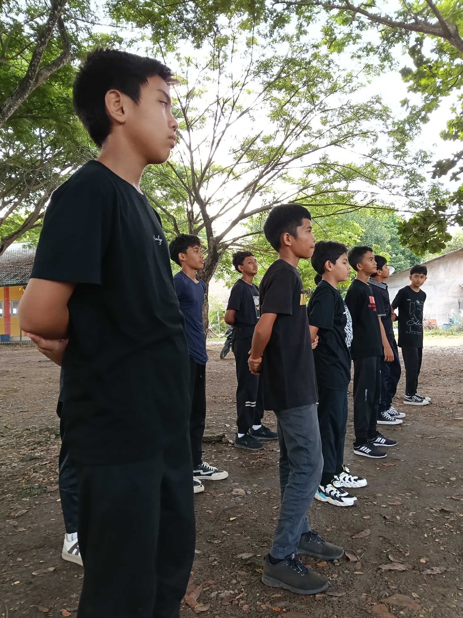 In Lhokseumawe, Indonesia, a group of boys stand in a line outdoors on a dirt-covered ground beneath trees. All are facing the same direction with their hands behind their backs. They vary in clothing; most wear black shirts and pants, but some wear navy or gray. The footwear ranges from sneakers to casual shoes. In the background are buildings, one with colored panels.