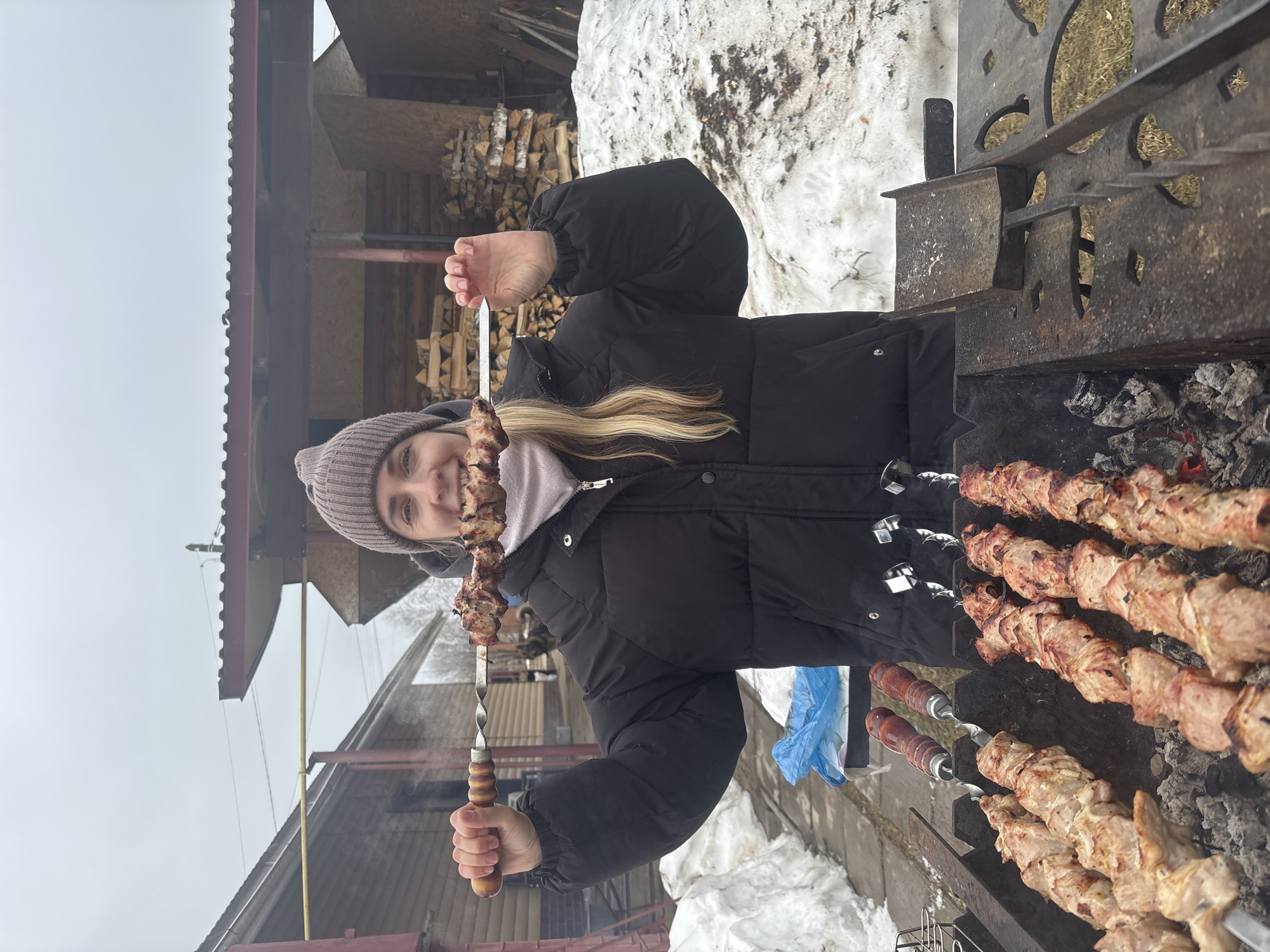 A young woman with blonde hair and a beanie is holding a skewer of grilled meat in front of her face. She is standing next to a grill with several skewers of meat cooking over hot coals. In the background, there is a building and snow on the ground. This photo was taken in Chaikovsky, Russia.