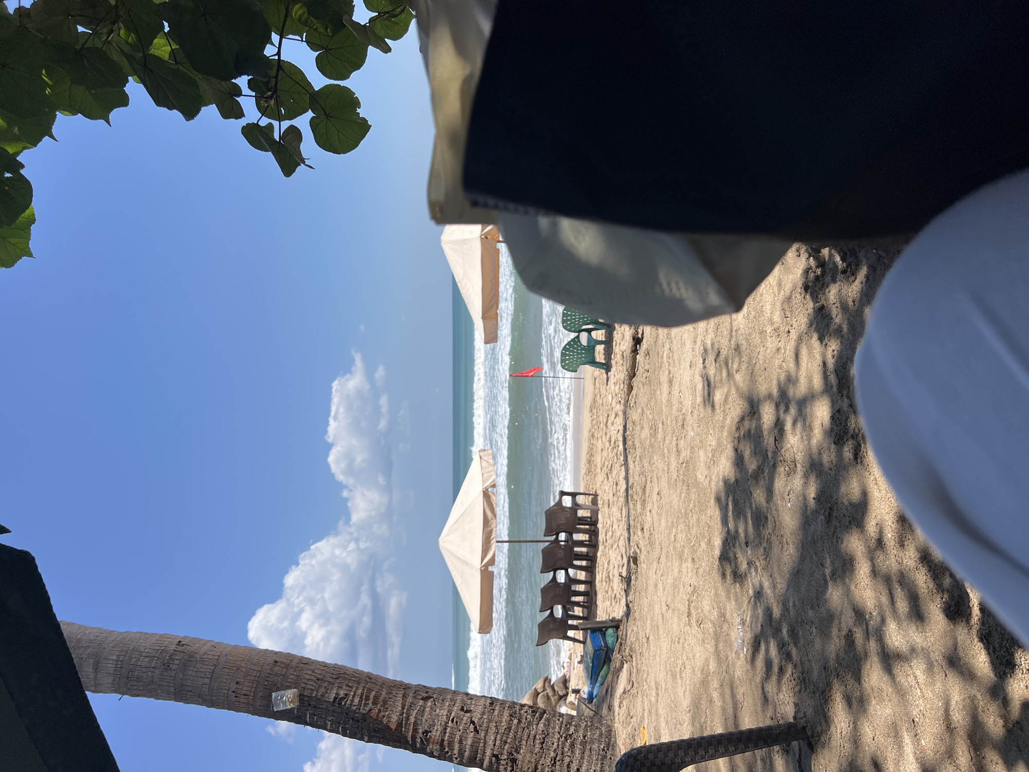 The image depicts a serene beach scene in Kuta, Indonesia. The foreground is dominated by the textured sand and shadows cast by unseen foliage, with a hint of what appears to be a dark object, possibly a chair or bag, on the right. Further back, a tall palm tree stands to the left, its rough bark textured and its trunk casting a shadow on the sand. In the middle ground, a row of four empty brown chairs sits beneath a beige umbrella on the sandy beach. Beyond the chairs, the turquoise ocean stretches to the horizon, with gentle waves rolling onto the shore under a clear blue sky dotted with fluffy white clouds. Two green chairs are visible closer to the water's edge, near a small red flag planted in the sand. A light brown umbrella stands behind the chairs. In the far background, there is a mountainous landscape. The overall impression is one of a tranquil and picturesque beachside setting.
