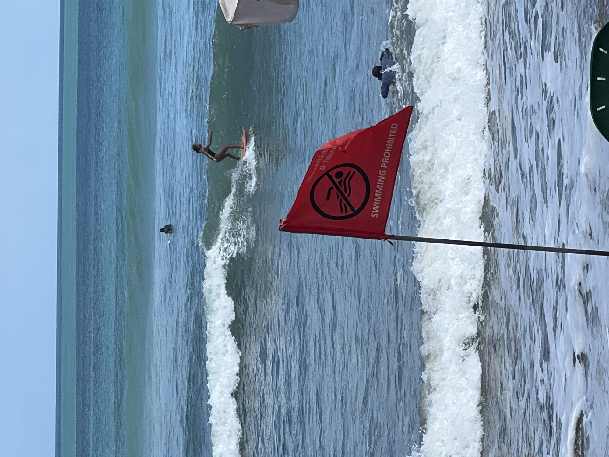 The image depicts a coastal scene in Kuta, Indonesia. A red flag with a circular "no swimming" symbol and the text "SWIMMING PROHIBITED" is prominently displayed in the foreground, planted on the beach. Beyond the flag, the ocean is active with moderate waves. In the water, a person is surfing on a red board, riding a wave. Further out, another person is visible in the water, appearing to be swimming or bodyboarding. The sky is clear blue, and the ocean exhibits shades of turquoise and deeper blue. The presence of the red flag indicates a potential hazard, likely strong currents or rough seas, making swimming unsafe despite the activity of surfers.