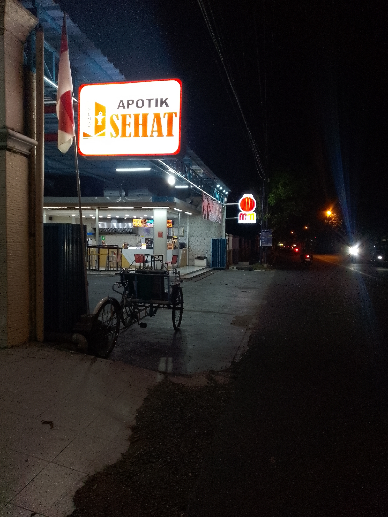 The image captures a nighttime scene outside an "Apotik Sehat" (Healthy Pharmacy) in Lamongan, Indonesia. A prominent illuminated sign for the pharmacy is visible, featuring text and a logo. To the left of the pharmacy entrance, an Indonesian flag is displayed. In front of the pharmacy, a three-wheeled vehicle, likely a pedicab or cart, is parked on the sidewalk. The pharmacy appears to be open, with lights on inside and visible kitchen equipment or serving counters. A street with a few vehicles and streetlights is located to the right of the pharmacy. The overall environment suggests a commercial area in Lamongan after dark.