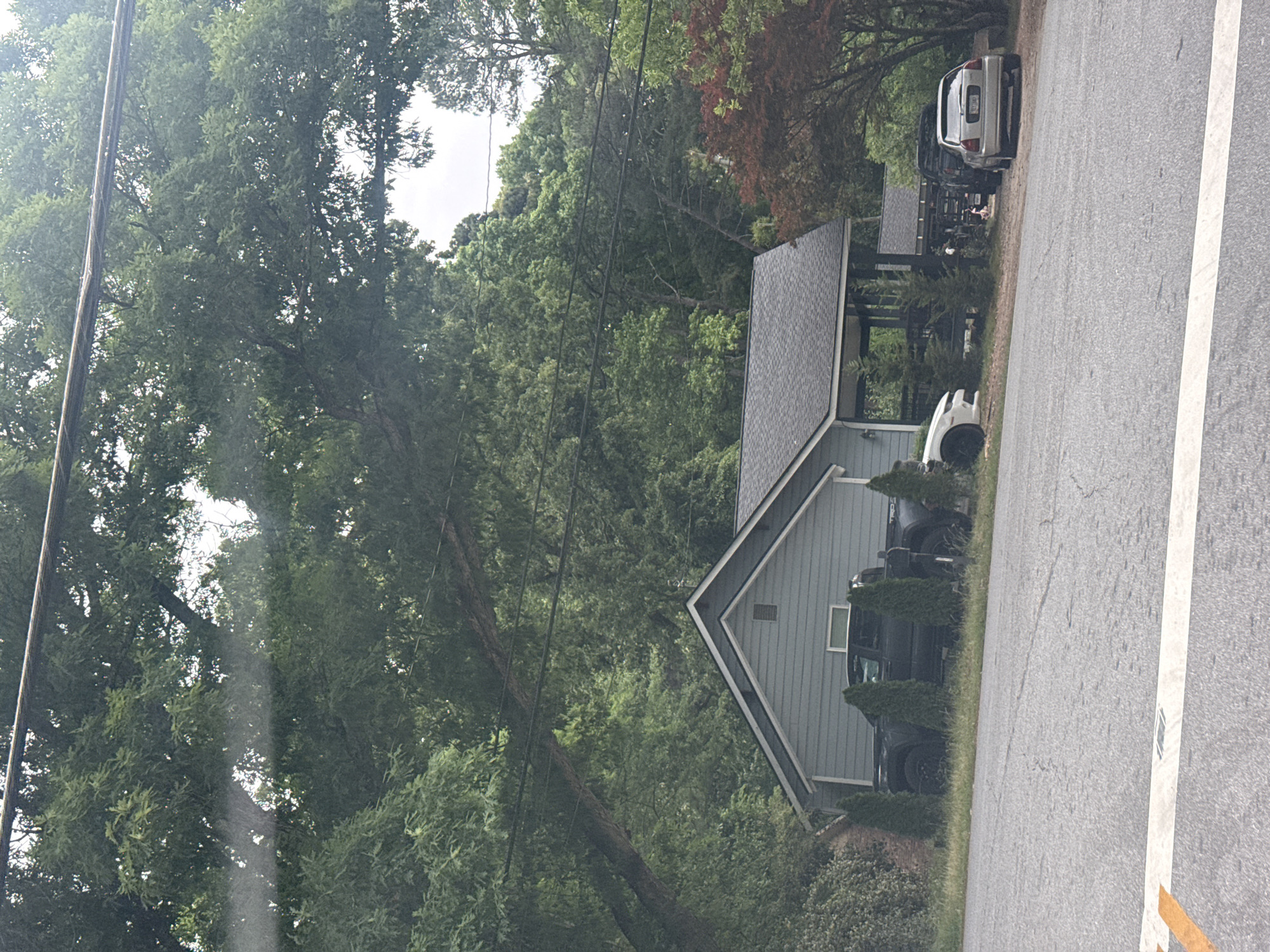 The image depicts a residential street in Mountain Park, Georgia, United States. A light blue, single-story house with a gray roof and a triangular front gable faces the road. Multiple vehicles are parked in the driveway and along the side of the road next to the house, including white, black and silver cars. The street is asphalt with a white line marking the edge. Abundant green trees and vegetation surround the house and line the street. Power lines are visible above the house and trees.
