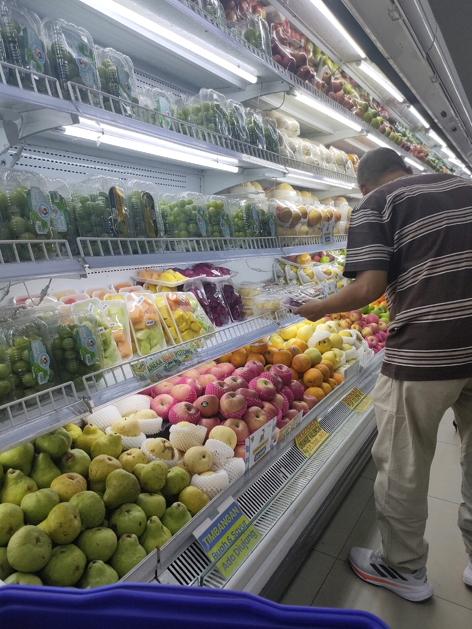 A middle-aged man with short, dark hair, wearing a brown and white striped t-shirt and light-colored pants, is browsing for fruit in a brightly lit supermarket. He is reaching for a package of grapes on a refrigerated display shelf. The shelves are stocked with a variety of fruits, including grapes in clear plastic containers, cut fruit salads in plastic trays, apples, pears, and oranges. The supermarket appears to be in Batam, Indonesia, as indicated by the Indonesian text on the price labels and signage, such as "TIMBANGAN Buah & Sayur" (Fruit & Vegetable Weighing). The flooring is tiled, and the overall environment is clean and well-organized.