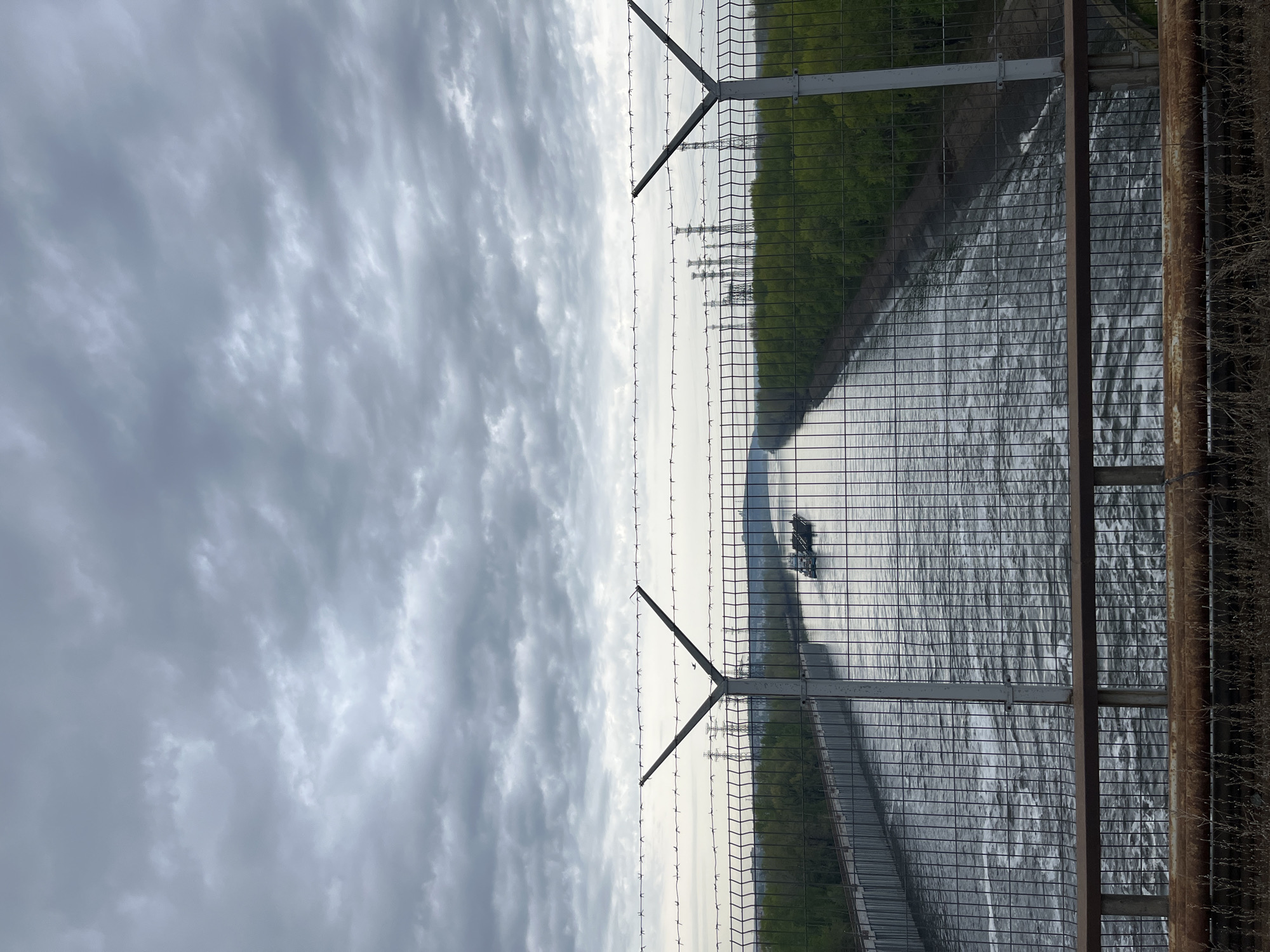 The image shows a view through a chain-link fence topped with barbed wire. In the background is a body of water, possibly a river or reservoir, with a green shoreline. Power lines and pylons are visible across the water. The sky is overcast with heavy clouds. A figure, possibly a person on a boat, is visible on the water. The location is likely near the city of Chaikovsky, Russia, based on the image's context.