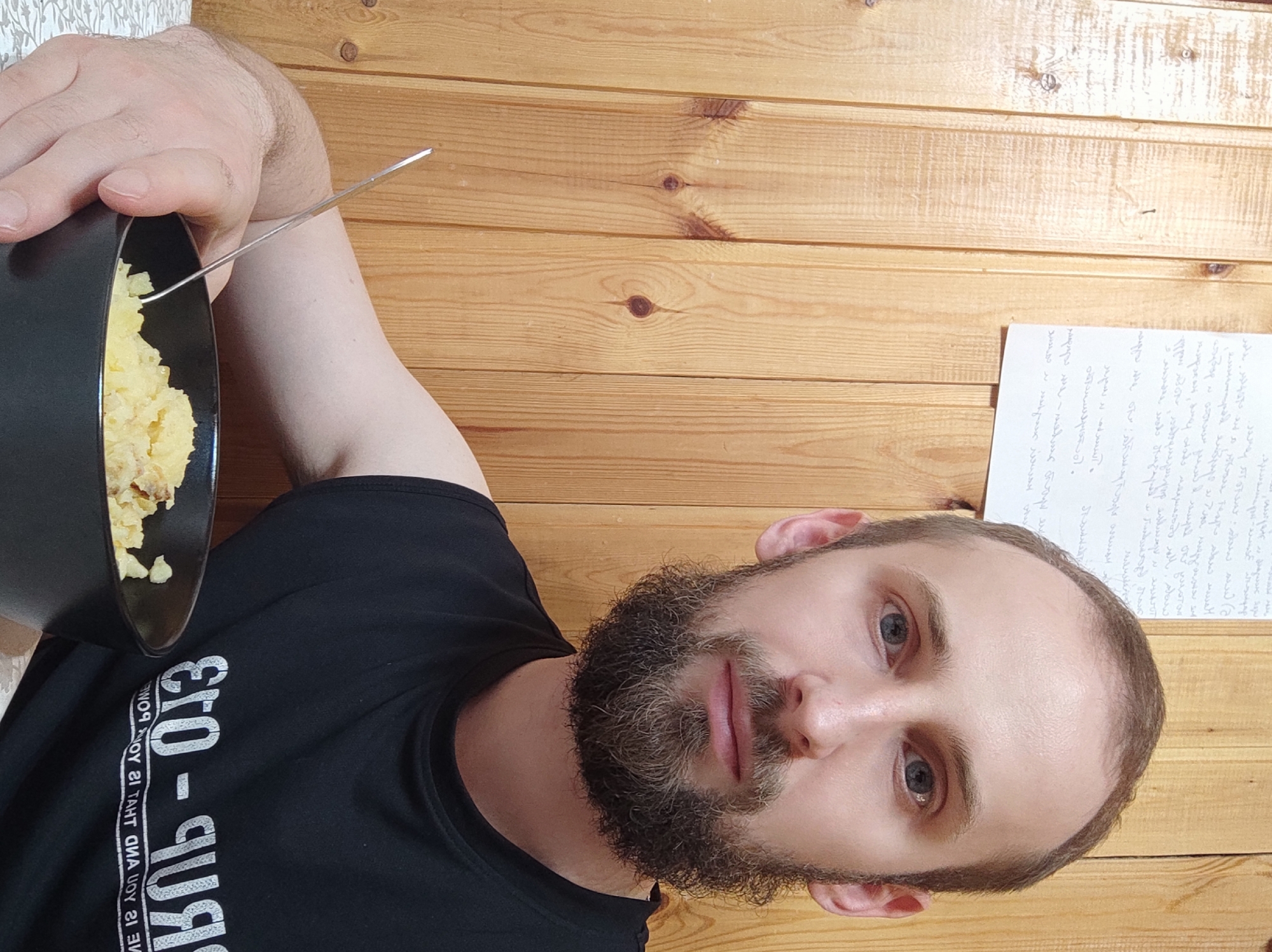 A man with a beard and gray eyes is holding a black bowl of food, possibly mashed potatoes, and a fork. He is wearing a black t-shirt with reversed white text. The background is a wooden wall, with a sheet of handwritten notes taped to it. The scene appears to be indoors.