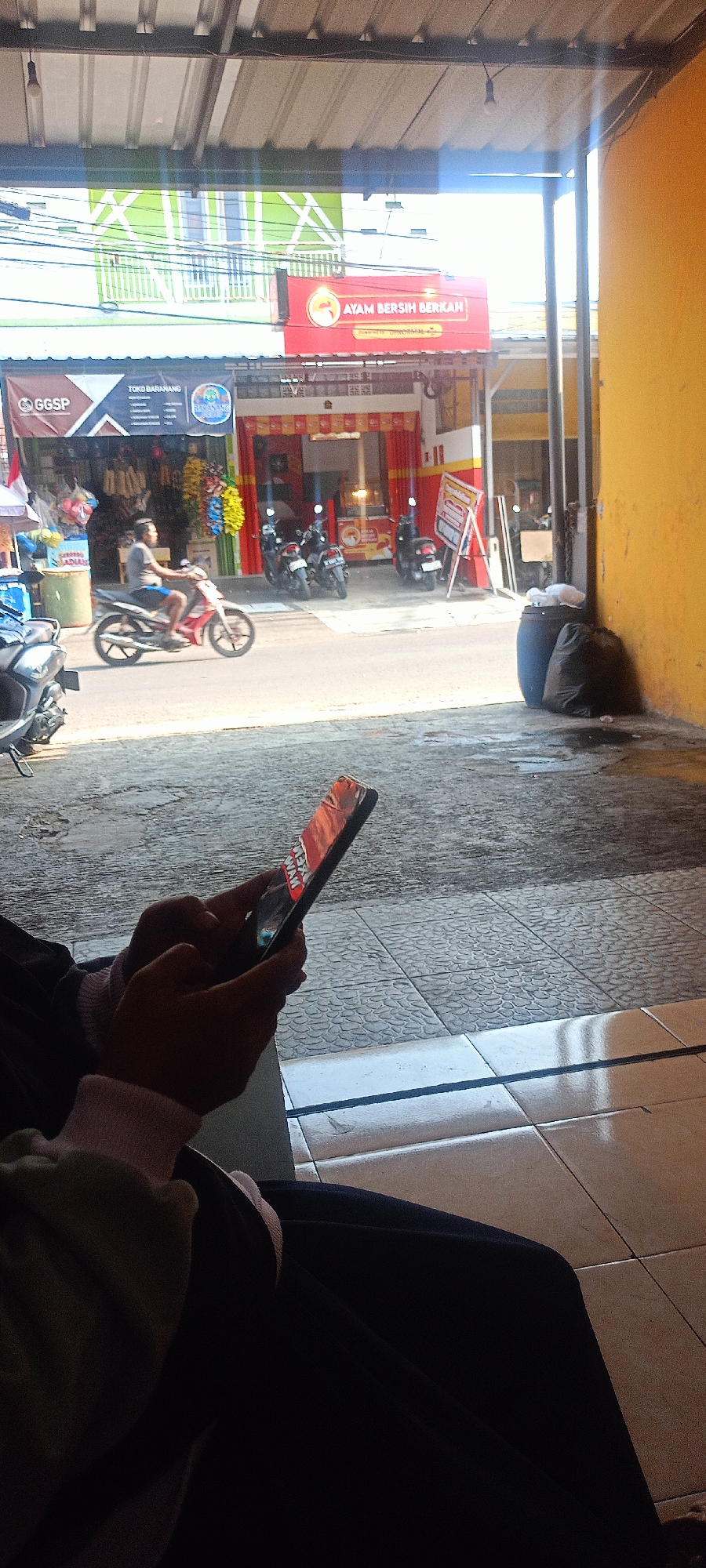 In Mekar Rahayu, Indonesia, a person is seated and holding a smartphone. The smartphone displays an image with text that reads "AYAM BERSIH BERKAH". In the background, there is a street scene with several motorcycles parked, and a man is riding a motorcycle. Buildings with various signage are visible, including one that says "TOKO BARANANG" and another with "GGSP". The environment appears to be a commercial area with shops and a street. The lighting suggests it is daytime.