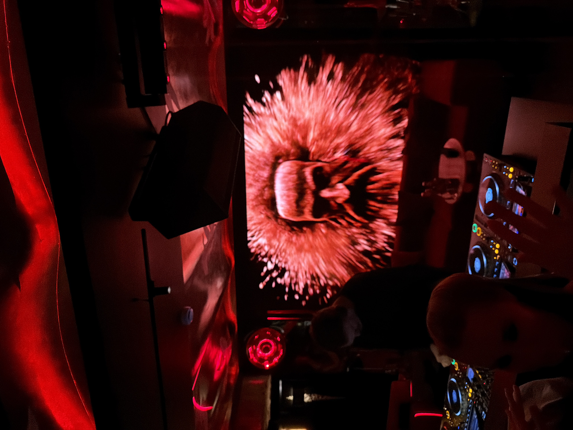 The image captures a dimly lit interior, bathed in red light. A screen displays an abstract image in shades of red and orange, resembling an explosion or a distorted face. To the left of the screen, a black speaker is visible. A DJ console with illuminated buttons and knobs is in the foreground, partially obscured by hands. The location is possibly in Moscow, Russia, but without further context, this cannot be verified.
