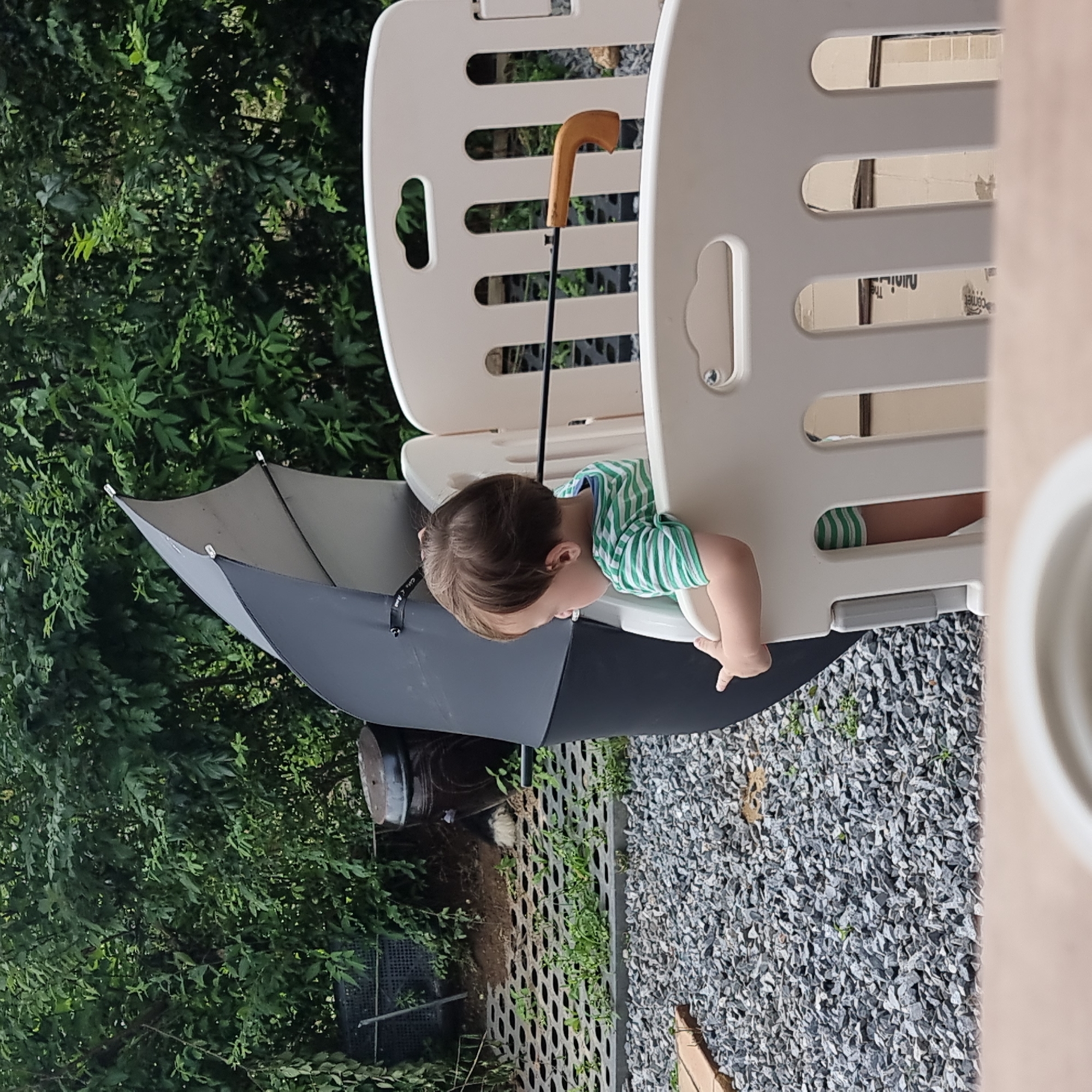 The image depicts an outdoor scene in Dunnae-myeon, South Korea. In the foreground, a baby is visible from the chest up, wearing a green and white striped shirt and green and white striped shorts. The baby is leaning against a white plastic playpen, holding onto its side. Behind the playpen, a large, partially opened black umbrella stands upright, with its wooden handle visible. The background consists of dense green foliage, indicating a natural or semi-natural environment. To the left of the umbrella, a large, dark, round pottery jar is situated next to a metal grate structure with plants growing through it. The ground is covered with small gray stones. The lighting suggests it is daytime, with diffused light, possibly indicating an overcast sky or shaded area.