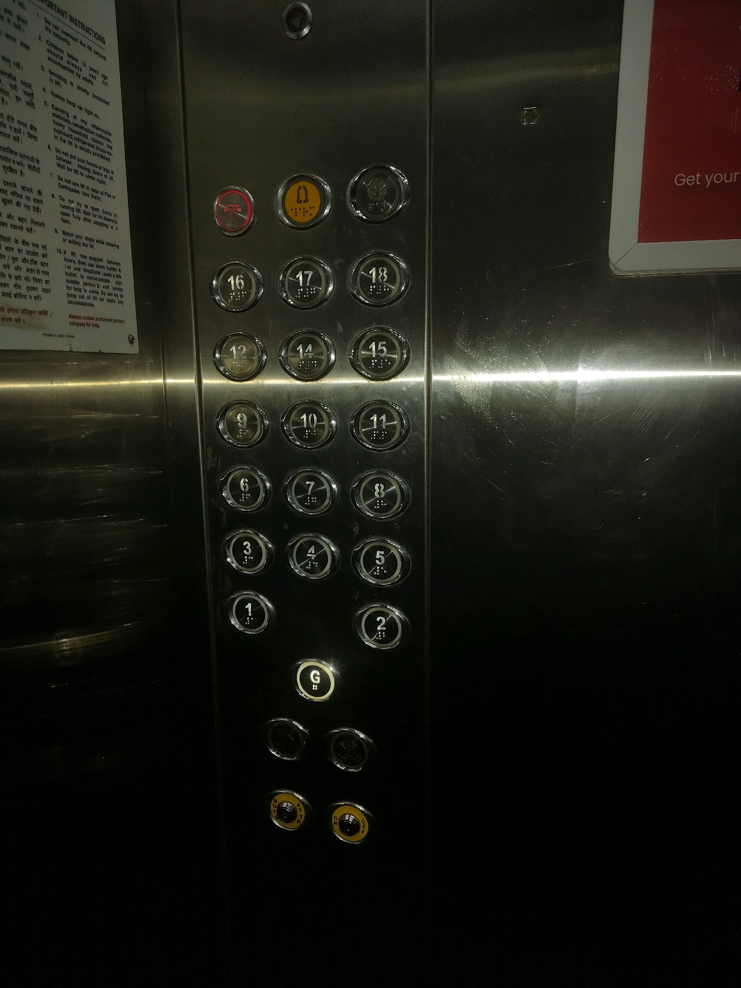 The image shows an elevator control panel, with a list of instructions in Hindi and English on the left side. The control panel is made of stainless steel and features several buttons, including floor numbers from G to 18, an alarm button, and two yellow buttons labeled "ATTENTION" and "ON/OFF." The elevator is likely located in a public building in Antarli, India, given the presence of Hindi language instructions and the typical design of an elevator panel in that region. The lighting in the image suggests it was taken indoors, possibly in a dimly lit hallway or lobby.