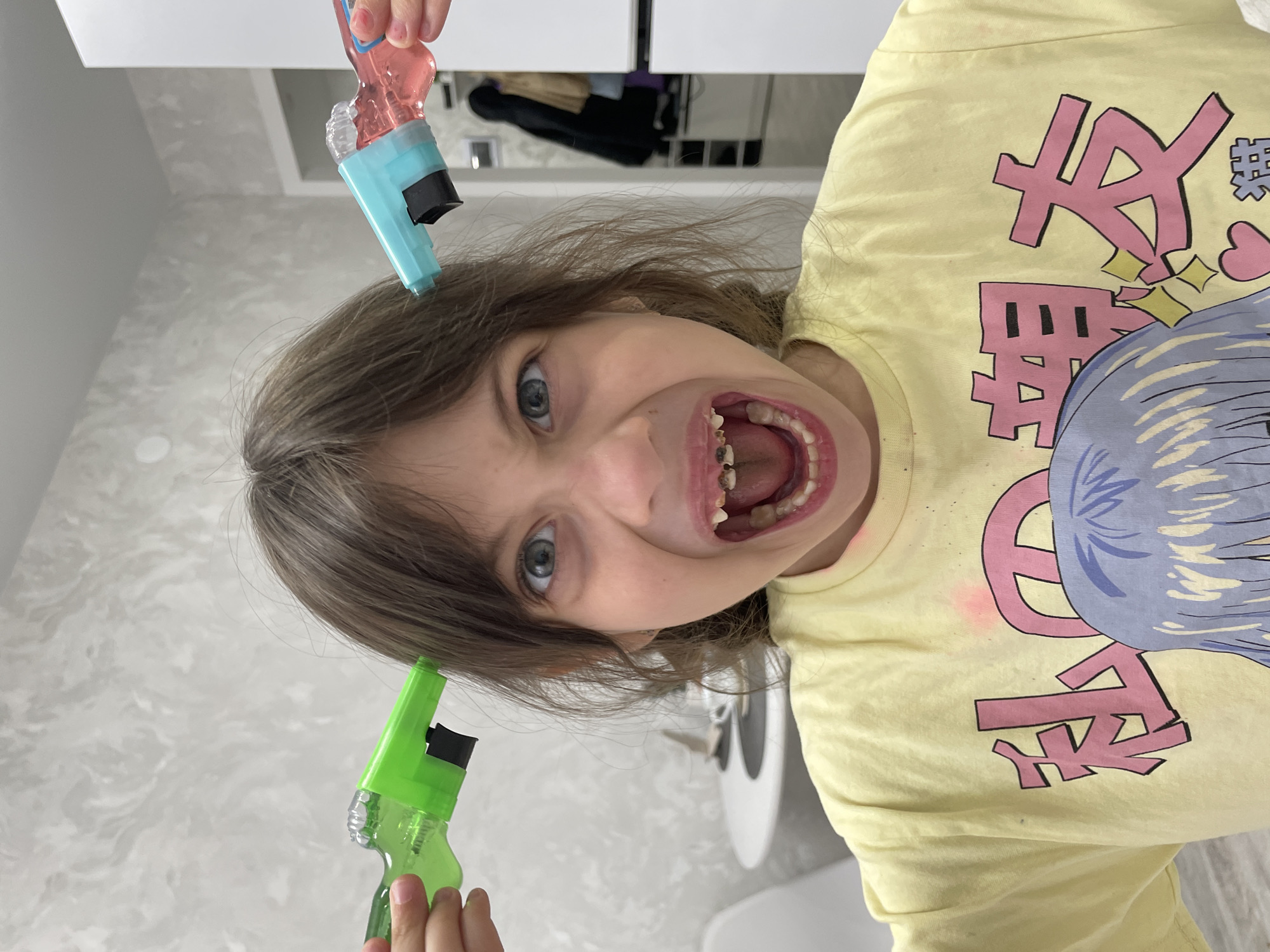 A young child with light brown hair and wide blue eyes is depicted indoors, looking directly at the camera with a wide-open mouth, revealing missing and dark-colored primary teeth. Her expression appears playful and mischievous. Two small, toy-like squirt guns are held near her head by unseen hands; one is bright blue with pink liquid, and the other is vivid green.
The child is wearing a light yellow t-shirt featuring large pink Japanese characters, possibly in an anime or manga style. Visible characters include "ハ" and "無", and other stylized strokes. The background shows a light-colored, subtly textured wall and white cabinetry, suggesting a bathroom or utility room setting. The image captures a candid moment of childhood play and excitement. The original orientation of the image is rotated 90 degrees counter-clockwise. The location context is Izhevsk, Russia.