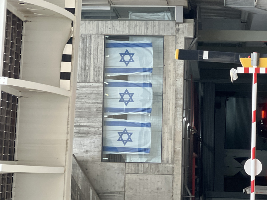 Three Israeli flags hang vertically within a glass enclosure set into a concrete wall. The wall features a raw, textured concrete finish. To the left, a white structure with dark windows and a black-and-white striped element are visible. To the right, a striped pole, a black boom, and a partially obscured doorway suggest a building or entryway. The overall impression is modern and architectural.
