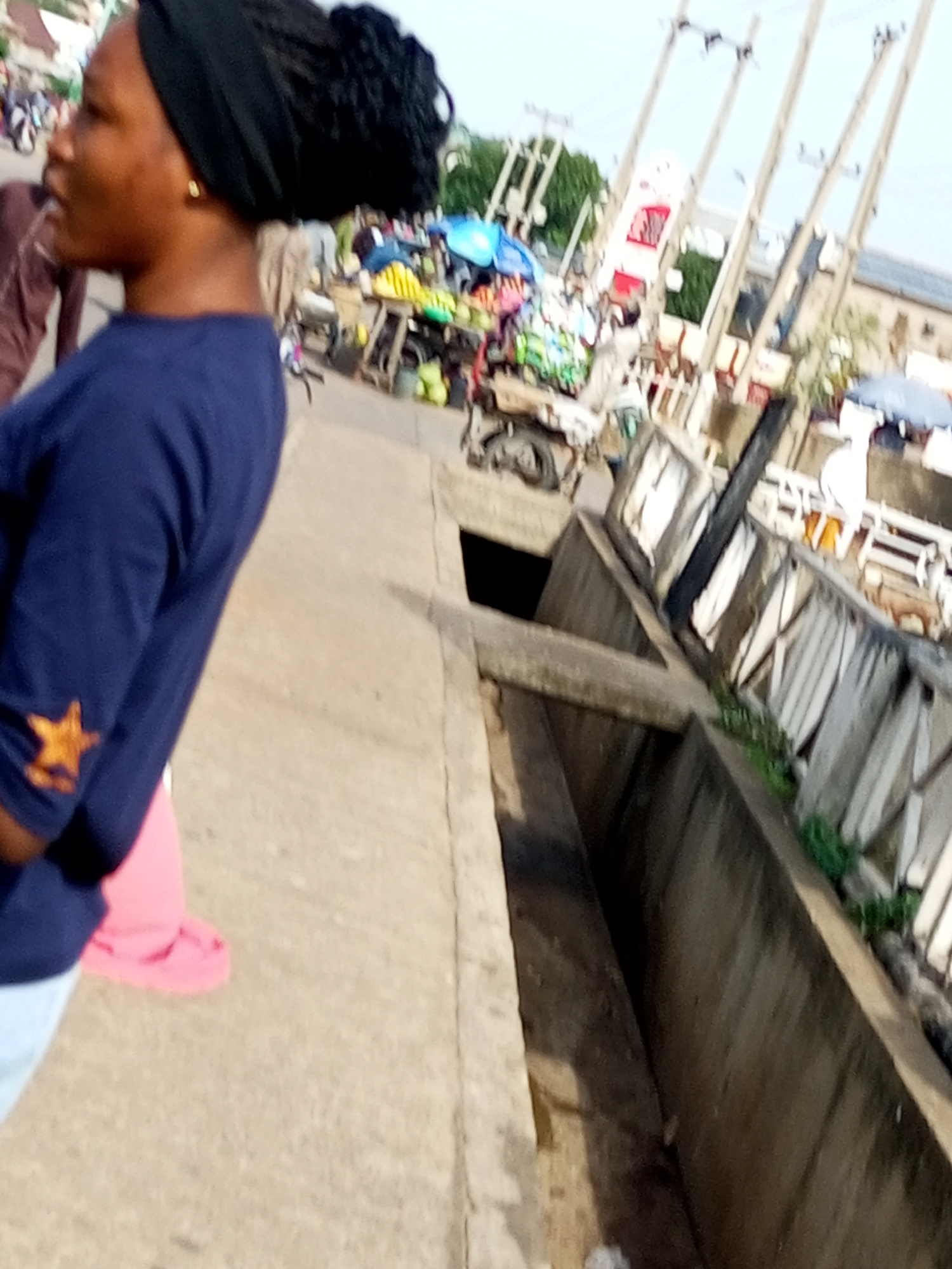 A close-up shot, captured in Tempure, Nigeria, focuses on the profile of a young Black woman from the chest up, facing left. She wears a dark blue long-sleeved shirt adorned with a small, embroidered orange star on the left sleeve. Her dark hair is braided and covered by a black headscarf. A single gold earring is visible in her left ear.
The background is a blur of activity, suggesting a bustling marketplace. Stalls with colorful goods, umbrellas, and what appear to be produce are visible. In the distance, several utility poles with wires crisscross the sky. A white and red structure, possibly a building or signage, is also present. The ground beneath the woman is paved with a textured material, possibly concrete or asphalt. To the right of the woman, a concrete drainage channel runs diagonally into the frame, its walls appearing weathered and damp. Some green vegetation is visible along the edge of the channel. The overall impression is a snapshot of daily life in an outdoor urban environment.